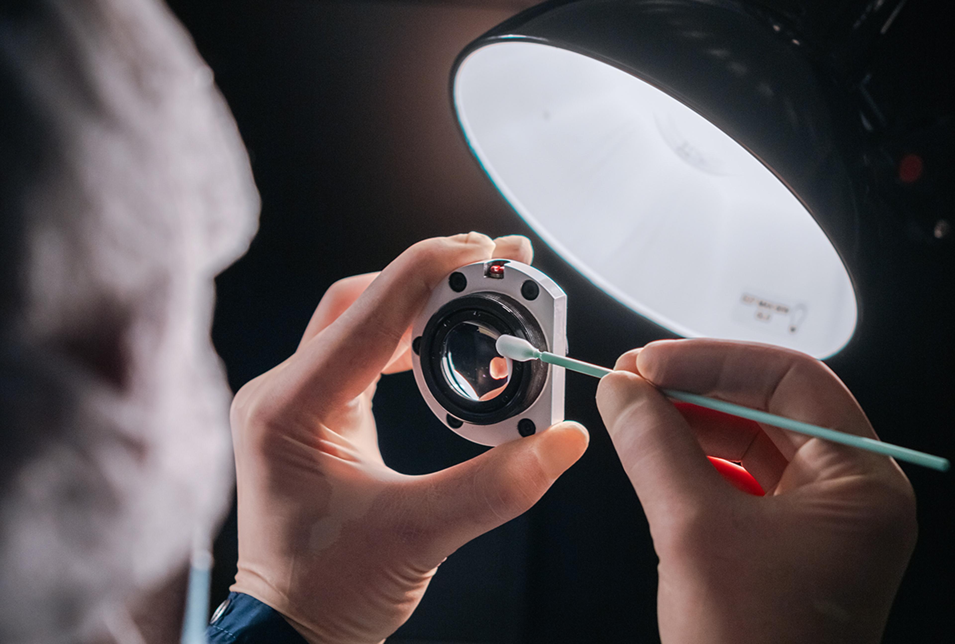 Close-up of a G&H technician carefully cleaning and inspecting a precision optical component under a bright lamp during optical assembly and quality control.