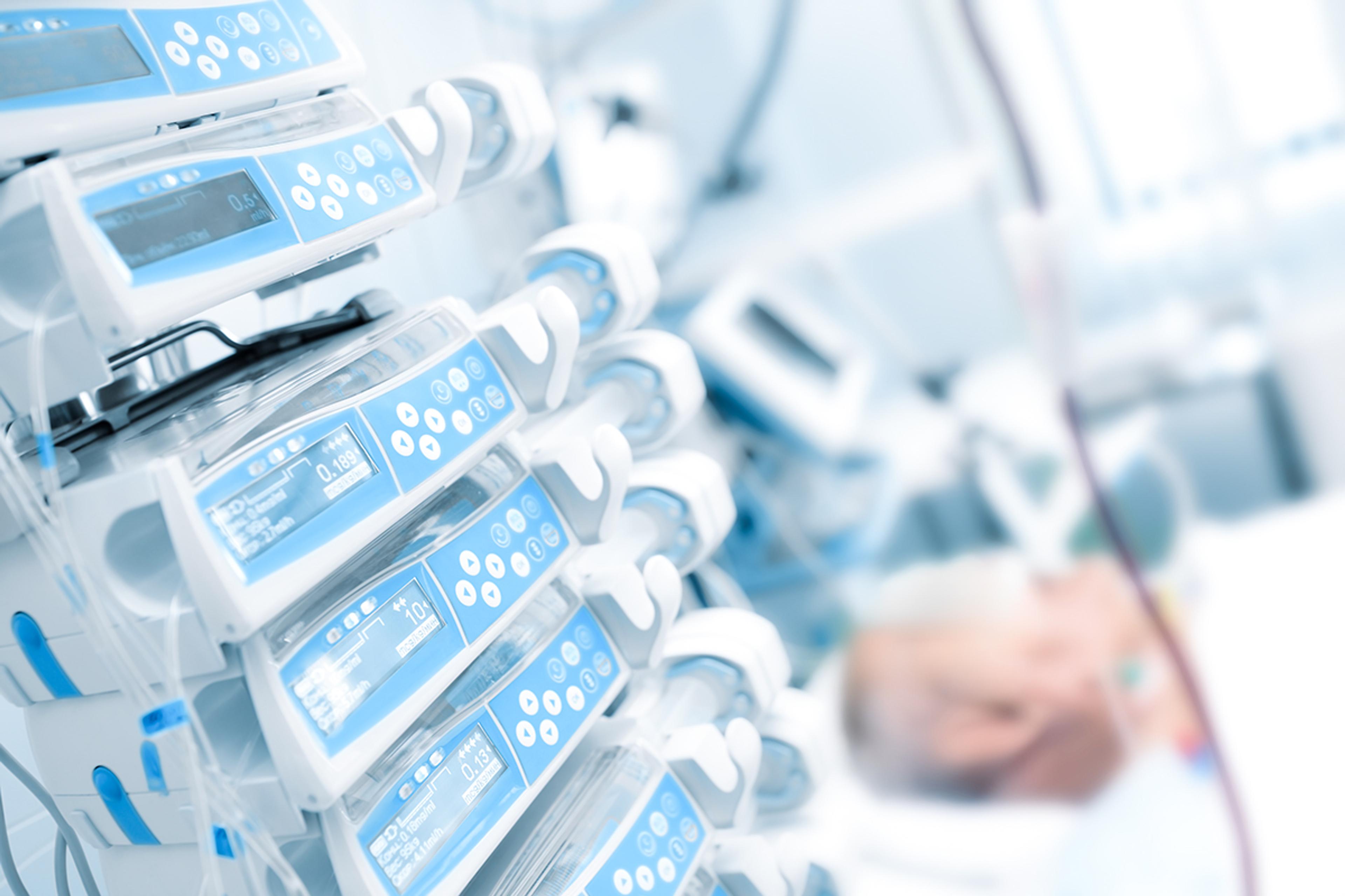 Close-up of multiple infusion pumps delivering controlled fluids or medication in a life-support hospital setting, with a patient resting blurred in the background, highlighting monitored care and clinical treatment support.