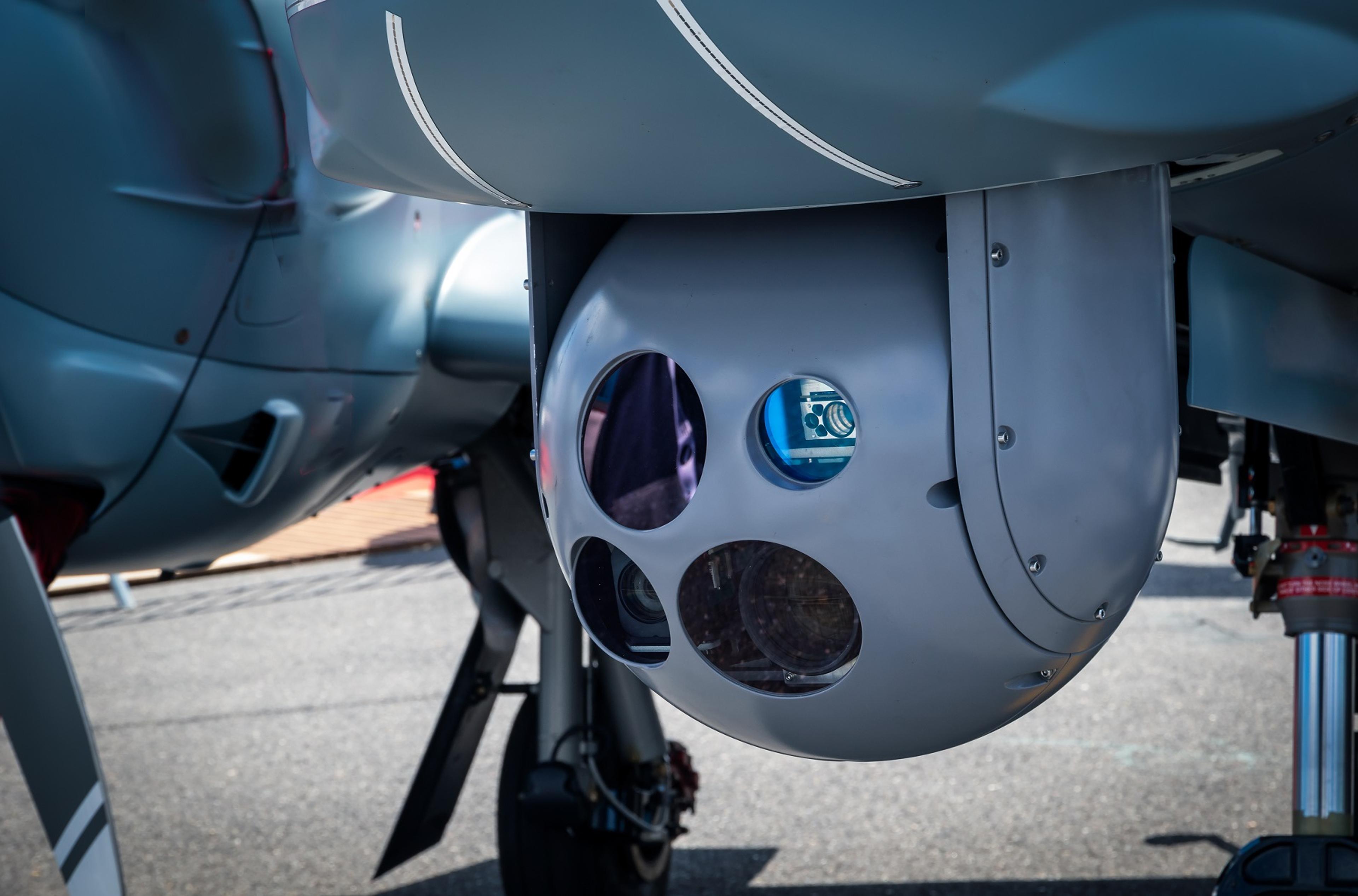 Gimbal - Close-up of a gimbal-mounted electro-optical/infrared (EO/IR) sensor system on the underside of an unmanned aerial vehicle (UAV). The sensor housing includes multiple optical windows, revealing internal lenses and electronic components used for surveillance and targeting applications. The aircraft is stationed on a tarmac.