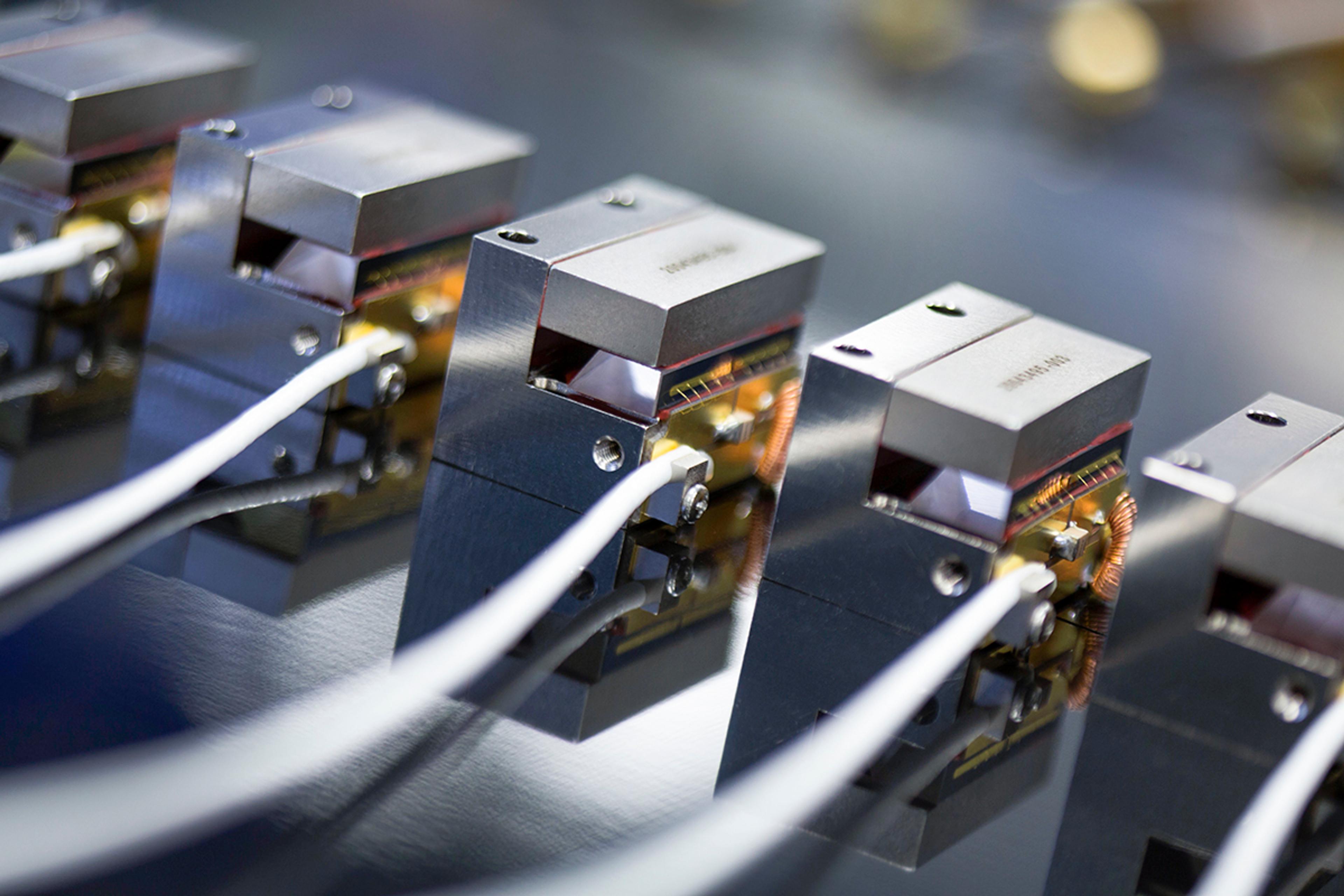 Close-up of G&H precision optical modules arranged in a row, with white fibre-optic cables connected to metal housings containing optical components and electronic coils for laser and photonics applications.