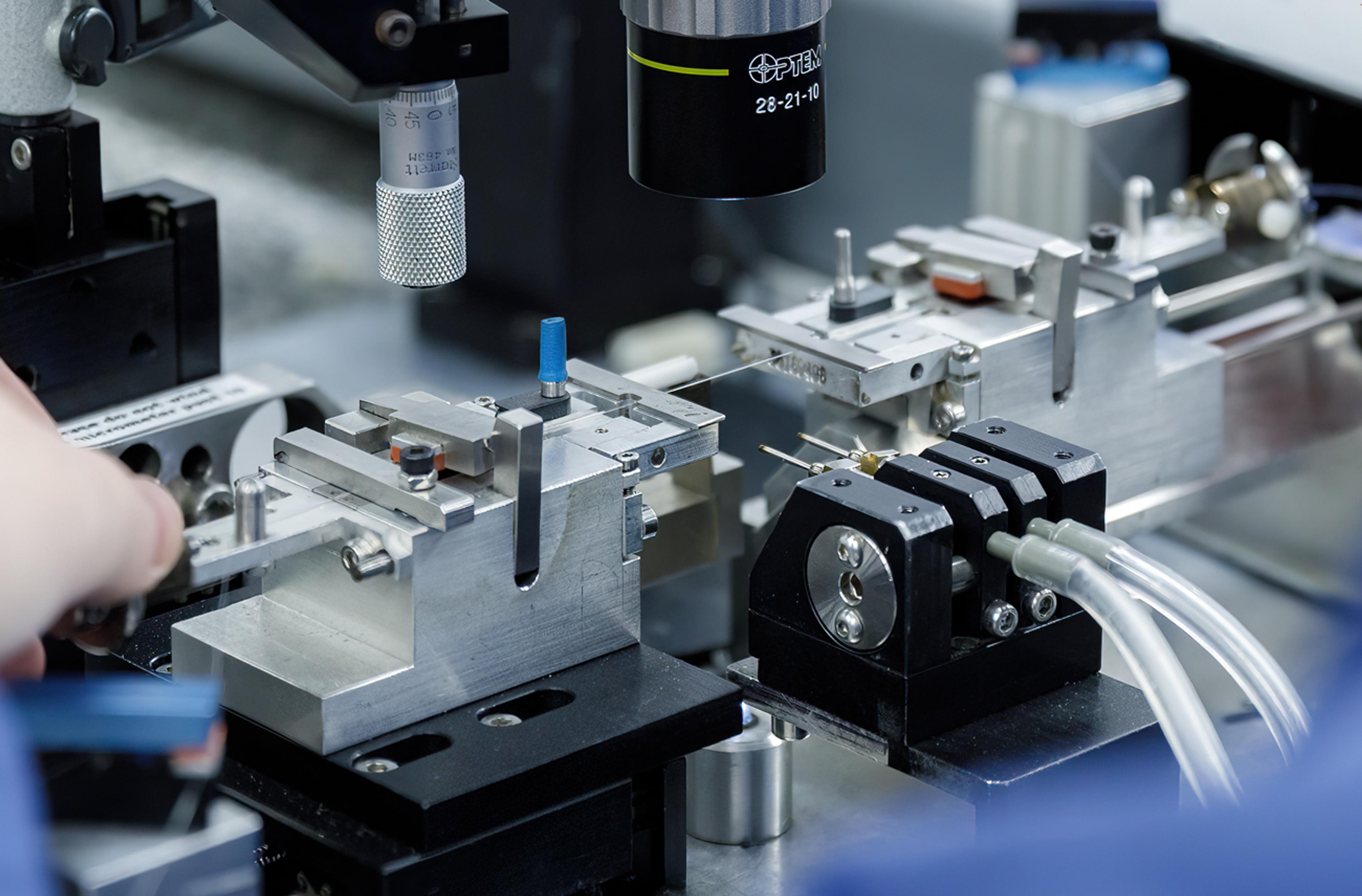 Close-up of a precision optical assembly featuring a fine fibre optic aligned between metal fixtures and micro-positioning stages, with tubing and a microscope objective used for accurate photonics component alignment and testing in a laboratory environment.