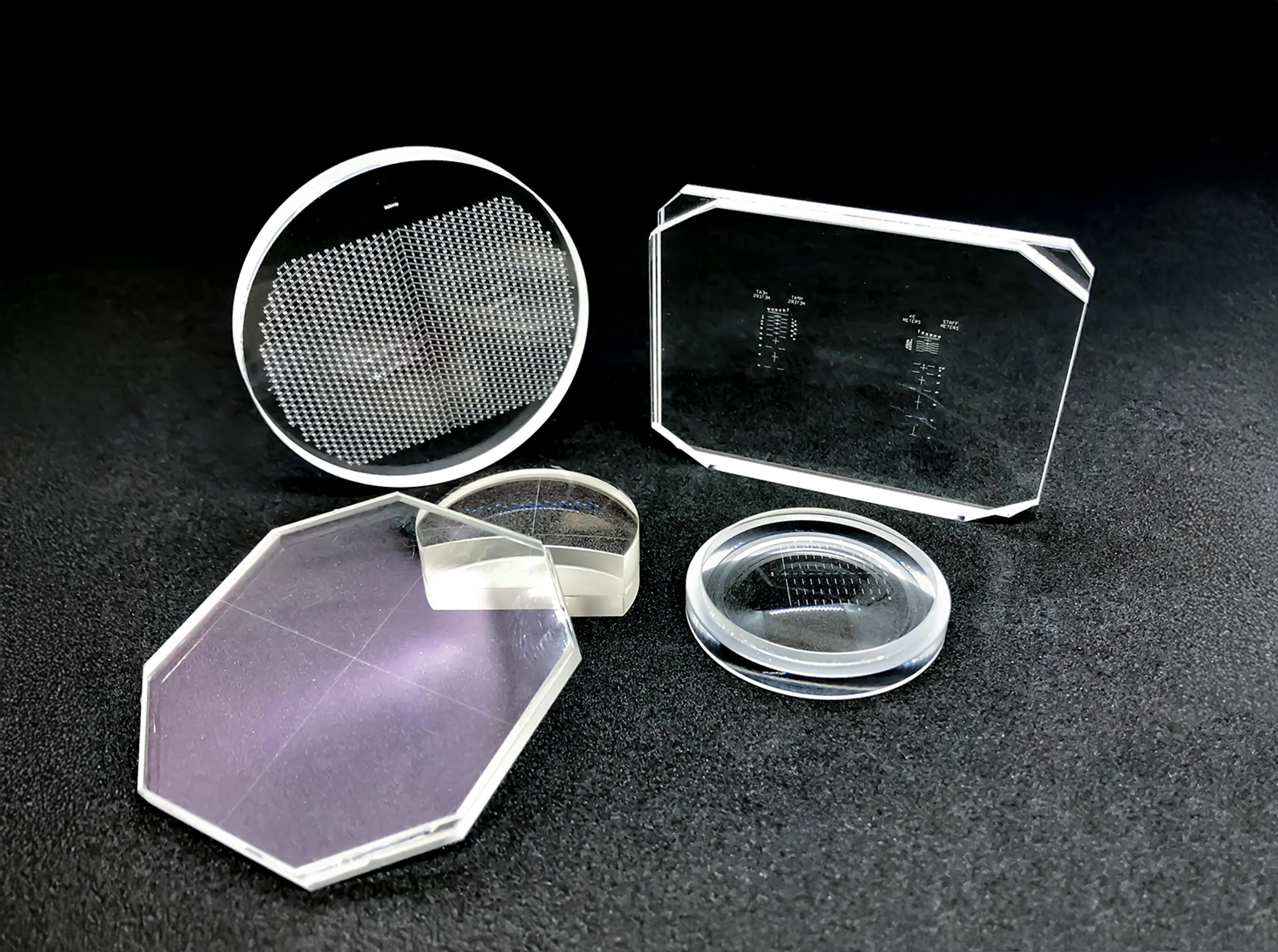A selection of optical reticles of various shapes and designs arranged on a dark surface. The set includes circular, rectangular, and octagonal glass components with etched patterns, mirrored surfaces, and transparent sections, used in precision optical applications.