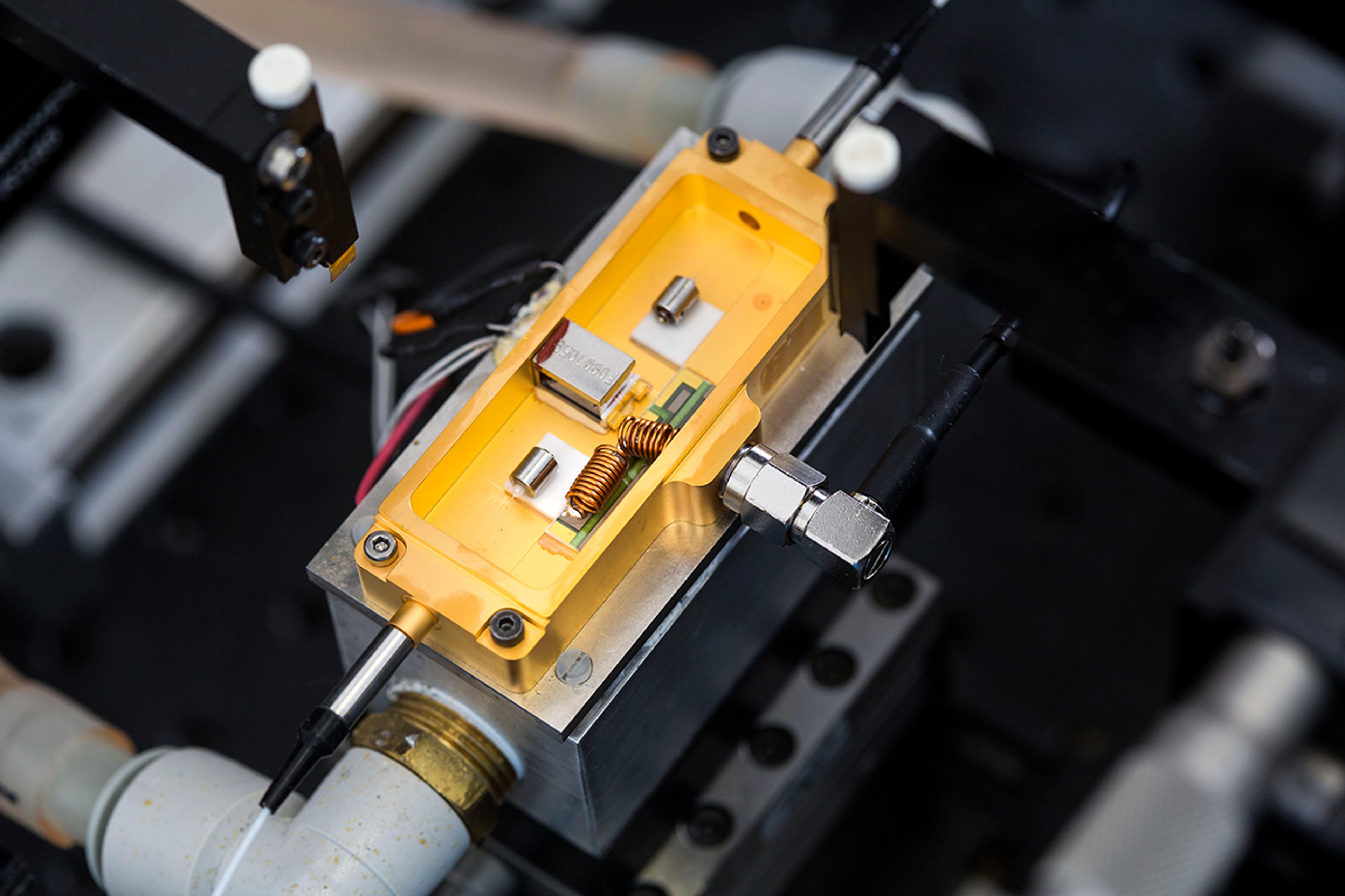 Close-up of an open gold fibre-coupled laser module on a test fixture, showing internal electronics and coil components with fibre and RF connections attached.
