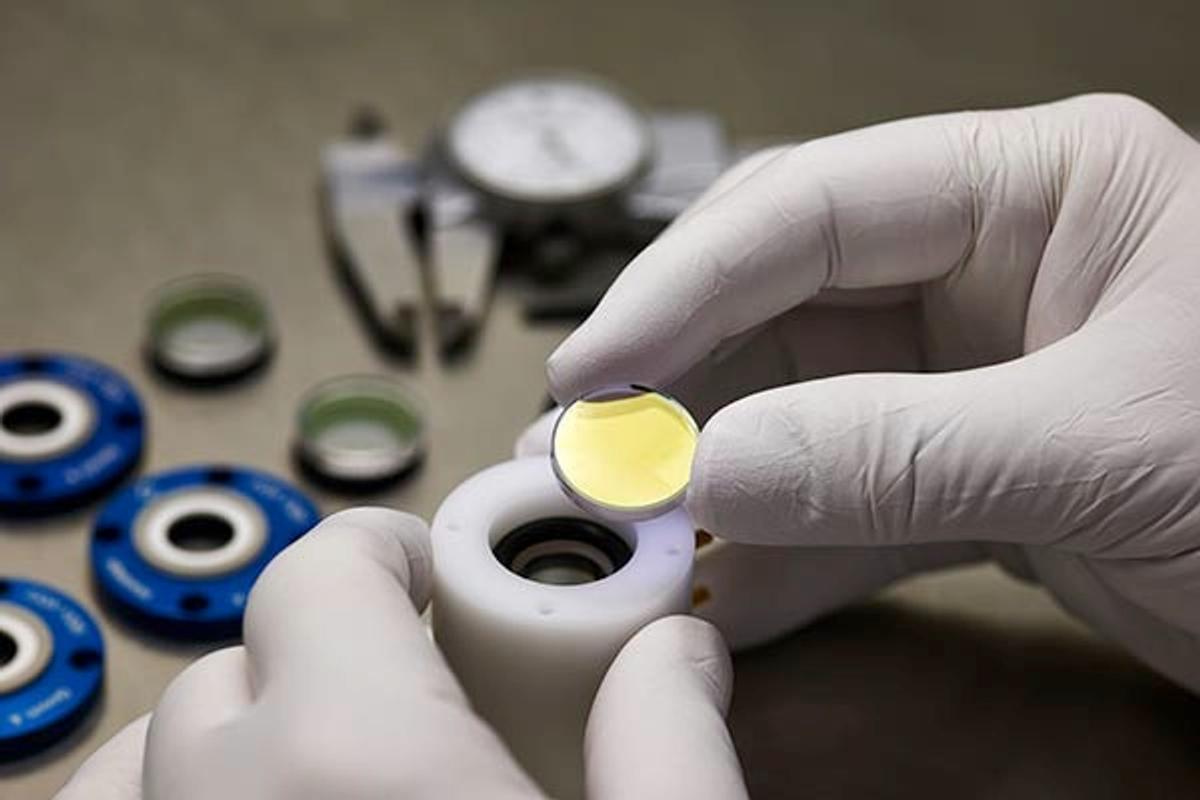 G&H pockels cells, A close-up image of a person wearing white gloves assembling or inspecting an optical component, likely a lens or crystal, with a yellow-tinted surface. The component is being placed into a white cylindrical holder. In the background, several other optical parts, including circular mounts and a precision measuring instrument, are visible on a workbench, indicating a high-precision optical or photonics assembly environment.