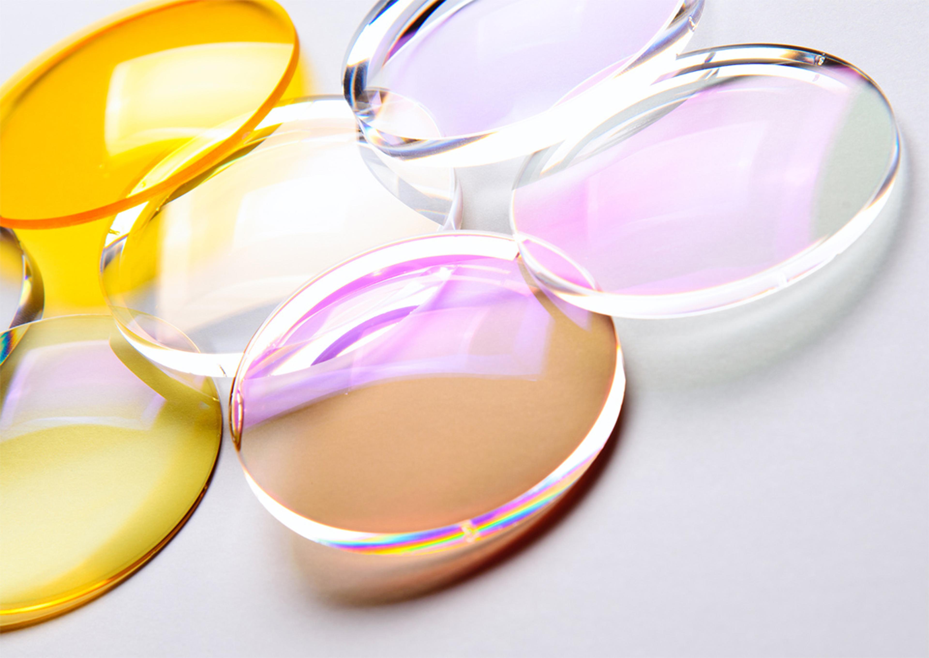 A close-up image of several precision polymer optical lenses in various hues, including yellow, clear, and pink-purple. The lenses are arranged on a light surface and exhibit subtle iridescent reflections, showcasing their high-quality optical coatings and transparency.