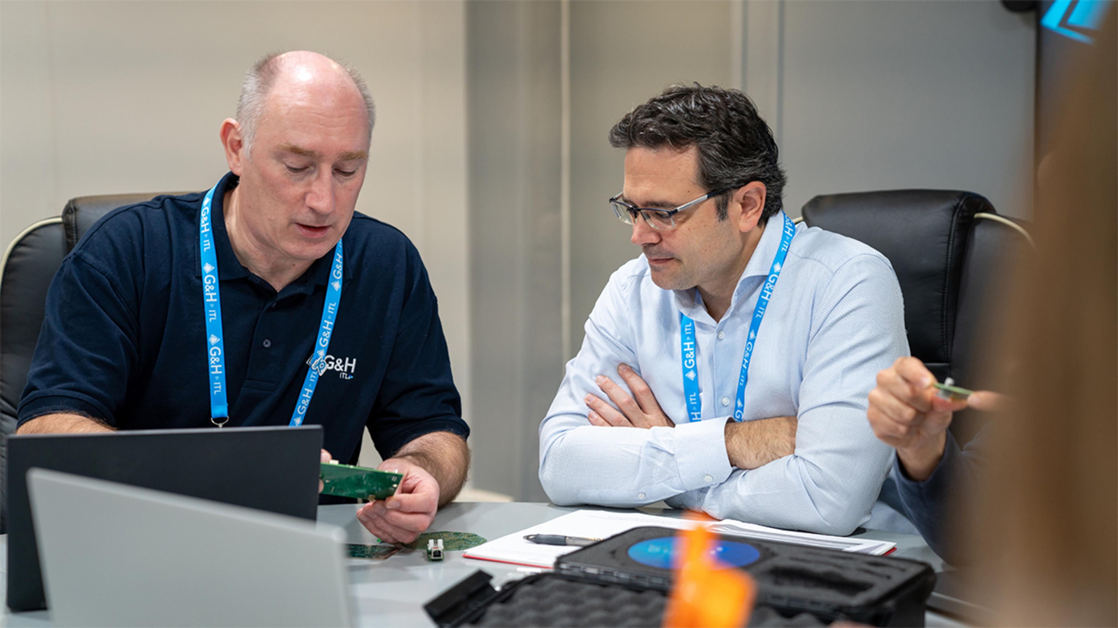 Two men wearing G&H ITL lanyards are seated at a conference table in a professional setting. One man, dressed in a navy blue G&H ITL polo shirt, is examining a green circuit board while explaining something to the other man, who is wearing glasses and a light blue shirt. Laptops, notebooks, and a case containing electronic components are spread across the table, indicating a technical or engineering discussion