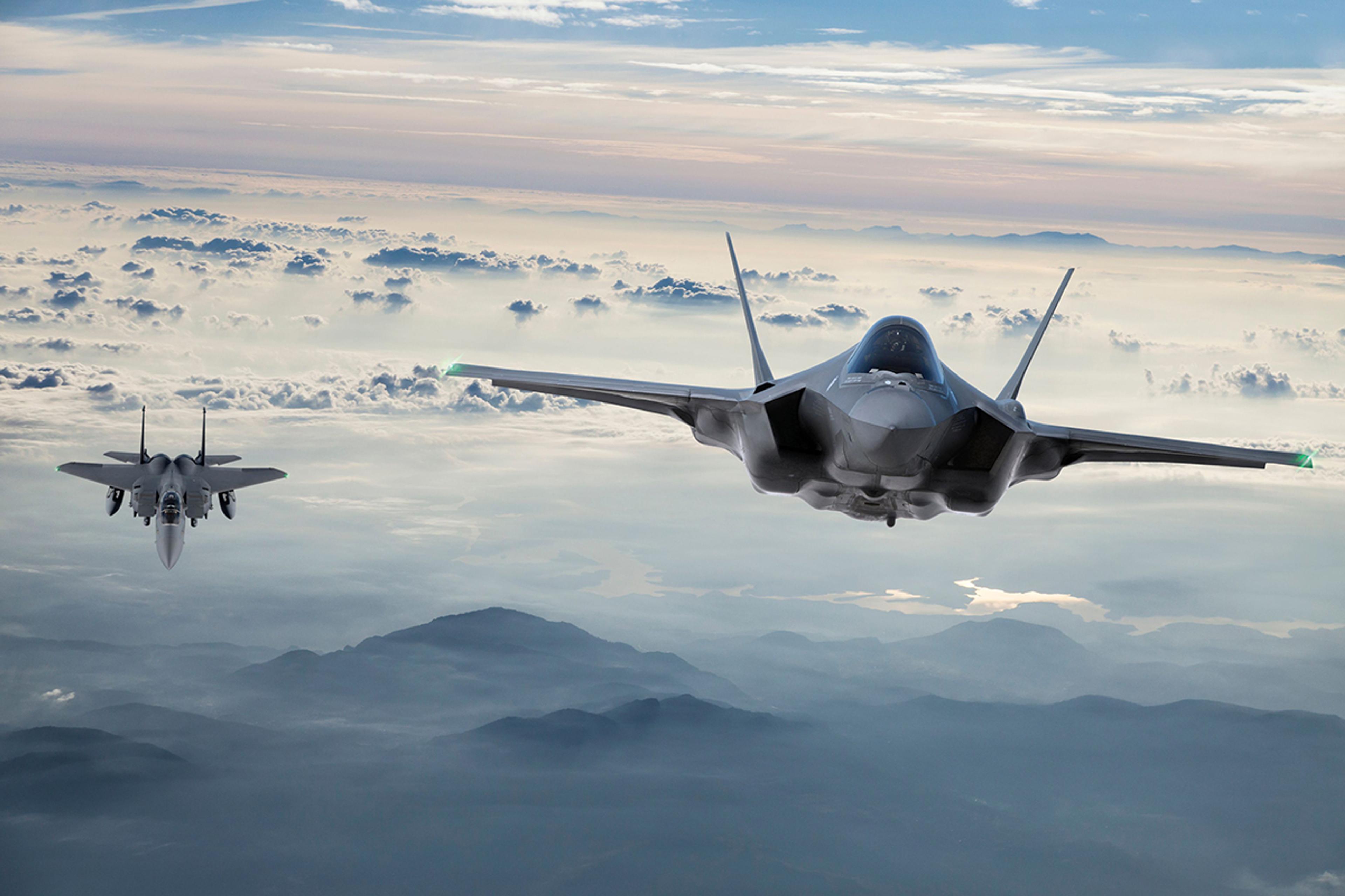 Two advanced military fighter jets flying above the clouds at high altitude, with mountainous terrain and a large body of water visible below, under a dramatic, partly cloudy sky at sunset.