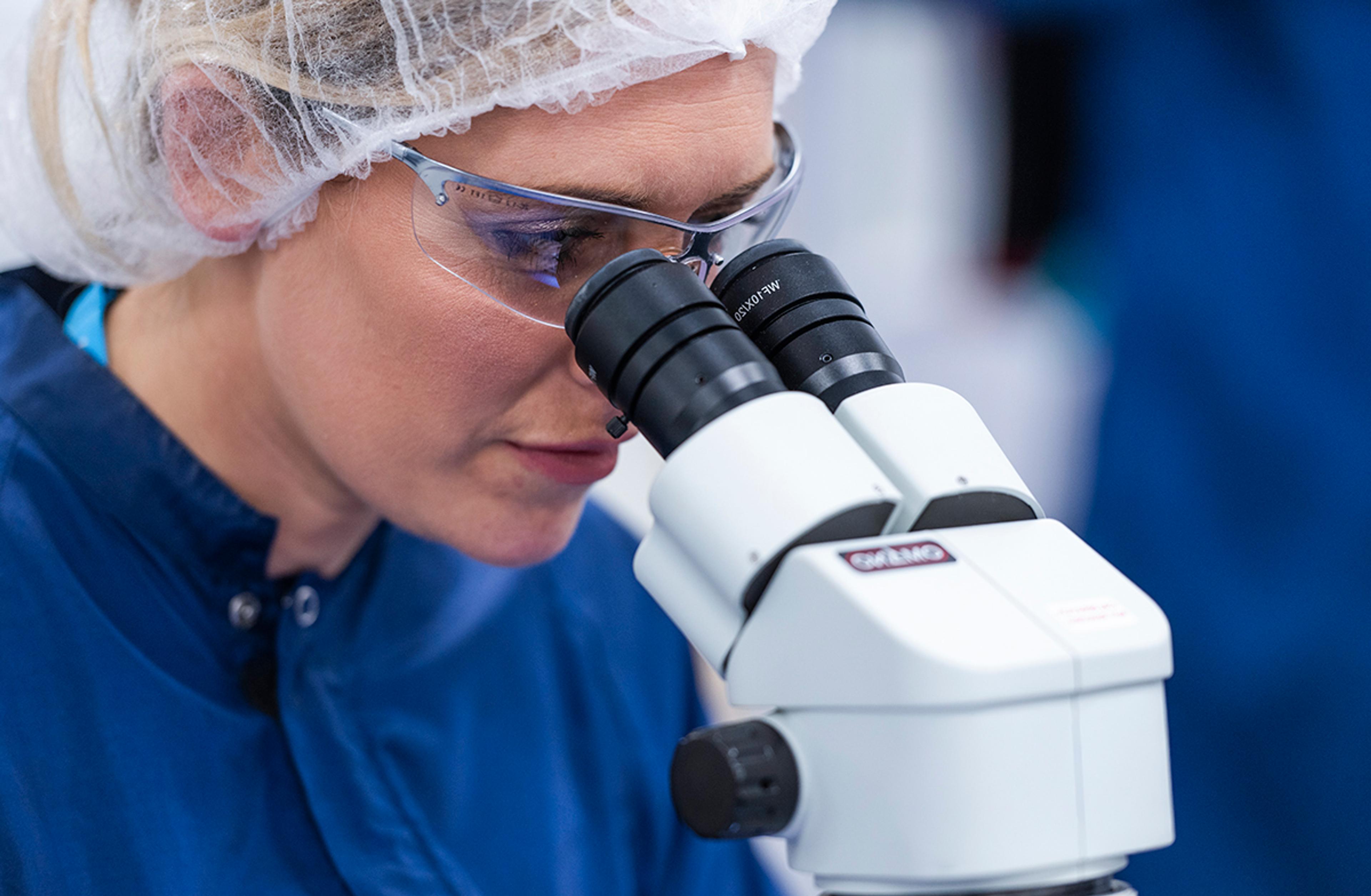 A woman wearing a hairnet, safety glasses, and a navy blue lab coat is closely examining something through a white binocular microscope in a laboratory setting. The background is softly blurred, emphasizing the scientific environment.