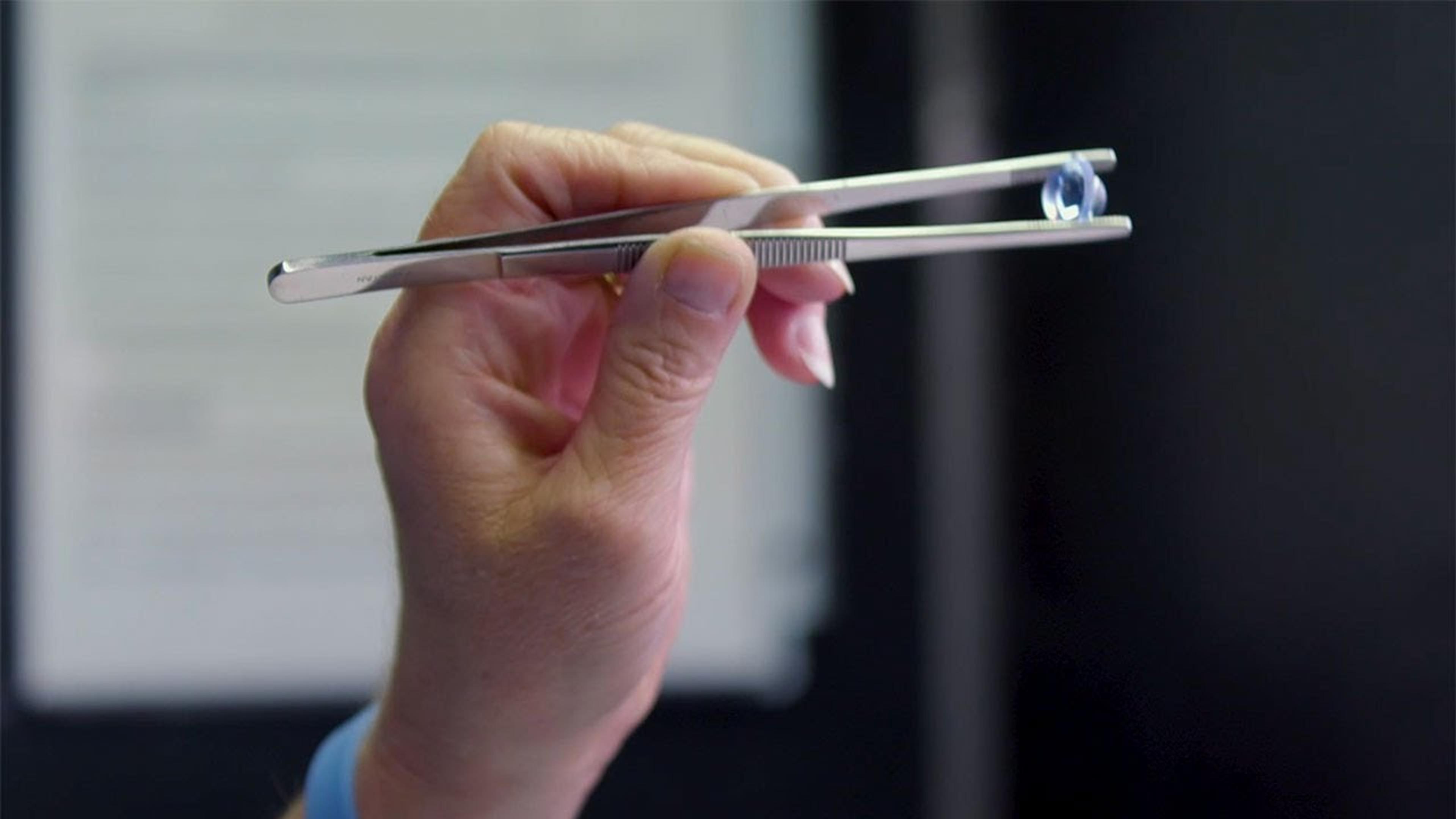 A hand holding precision tweezers that are gripping a small, round polymer optic lens. The lens appears clear with a slight blue tint, indicating optical coating. The background is softly blurred, focusing attention on the lens.