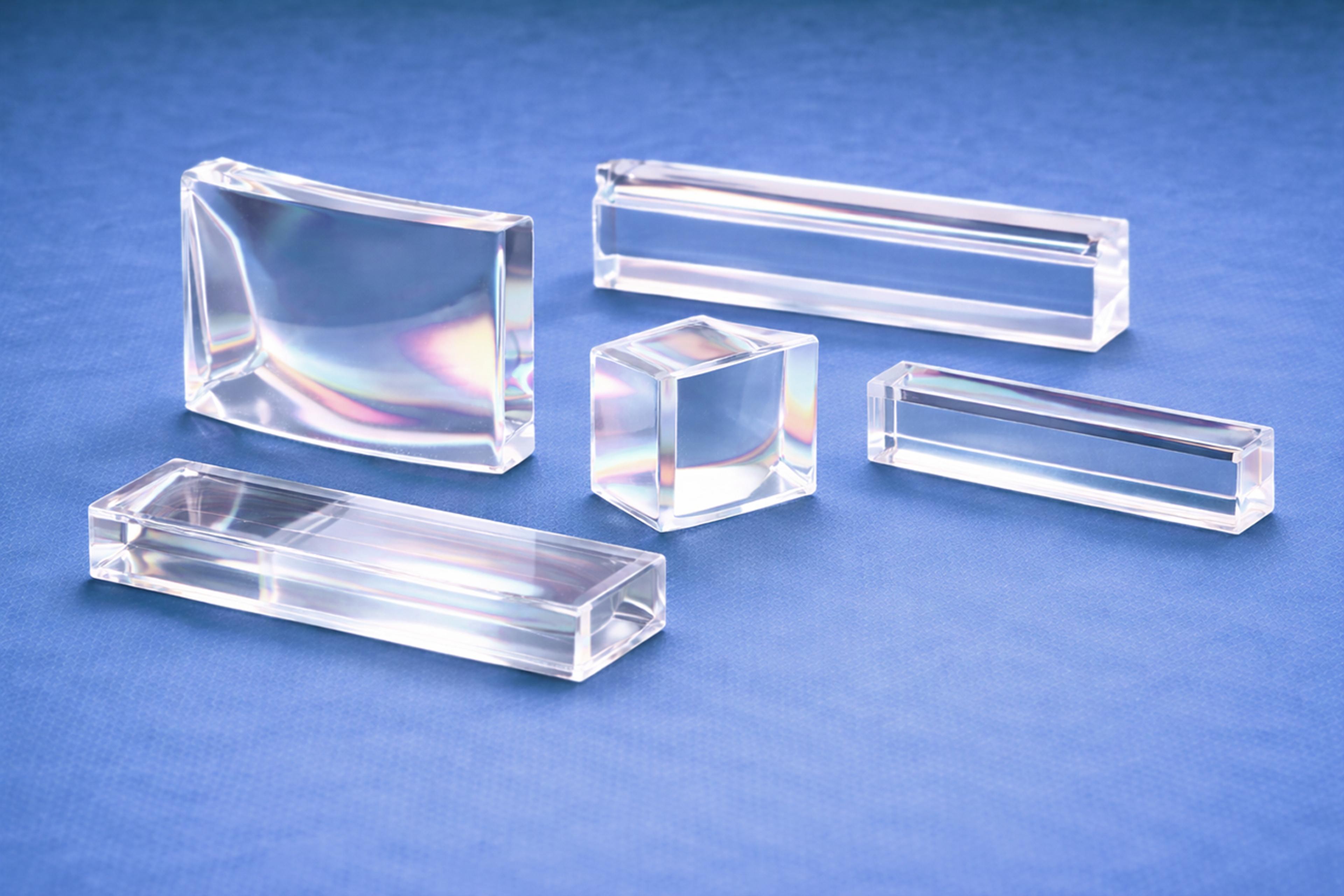 Transparent polymer beam splitter and prism optical components in rectangular and cubic forms, arranged on a blue background to illustrate precision polymer optics for beam steering and light management applications.