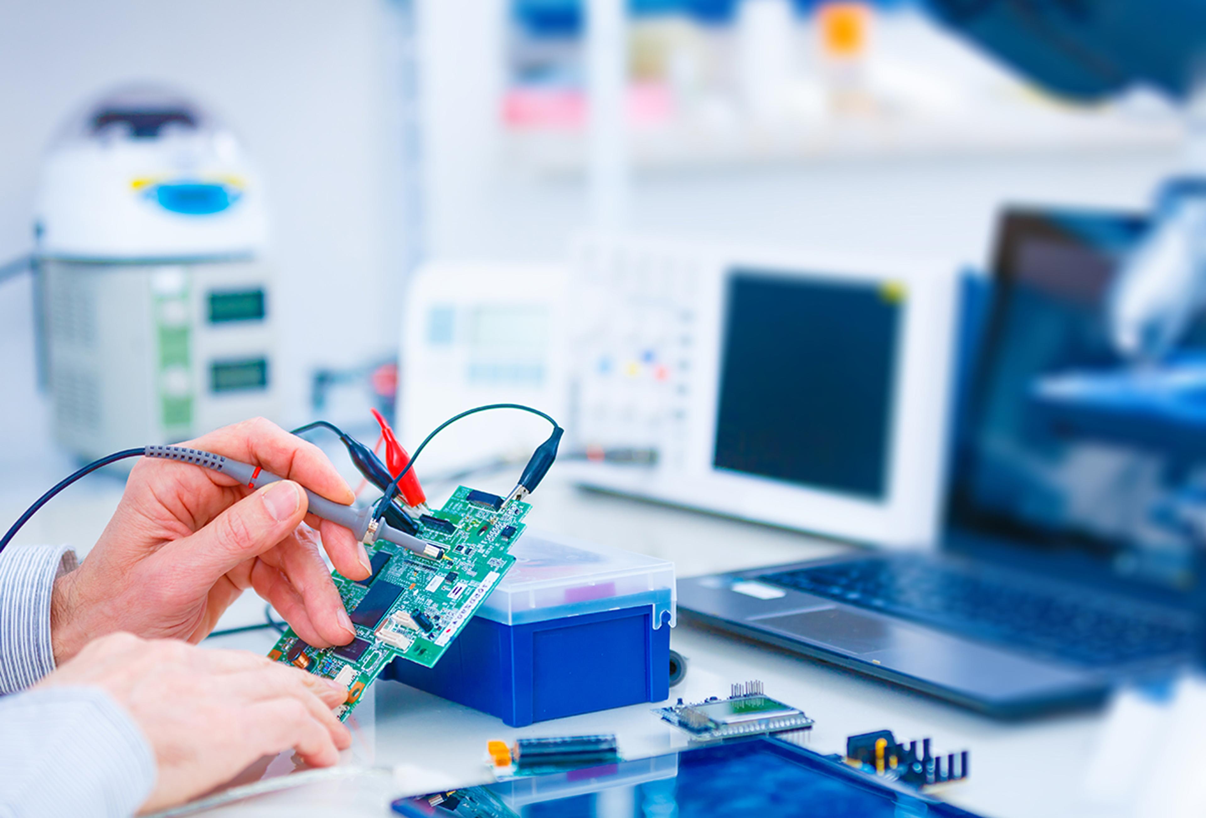 Engineer testing a green printed circuit board with probe leads and crocodile clips at an electronics lab bench, with instruments and a laptop in the background.