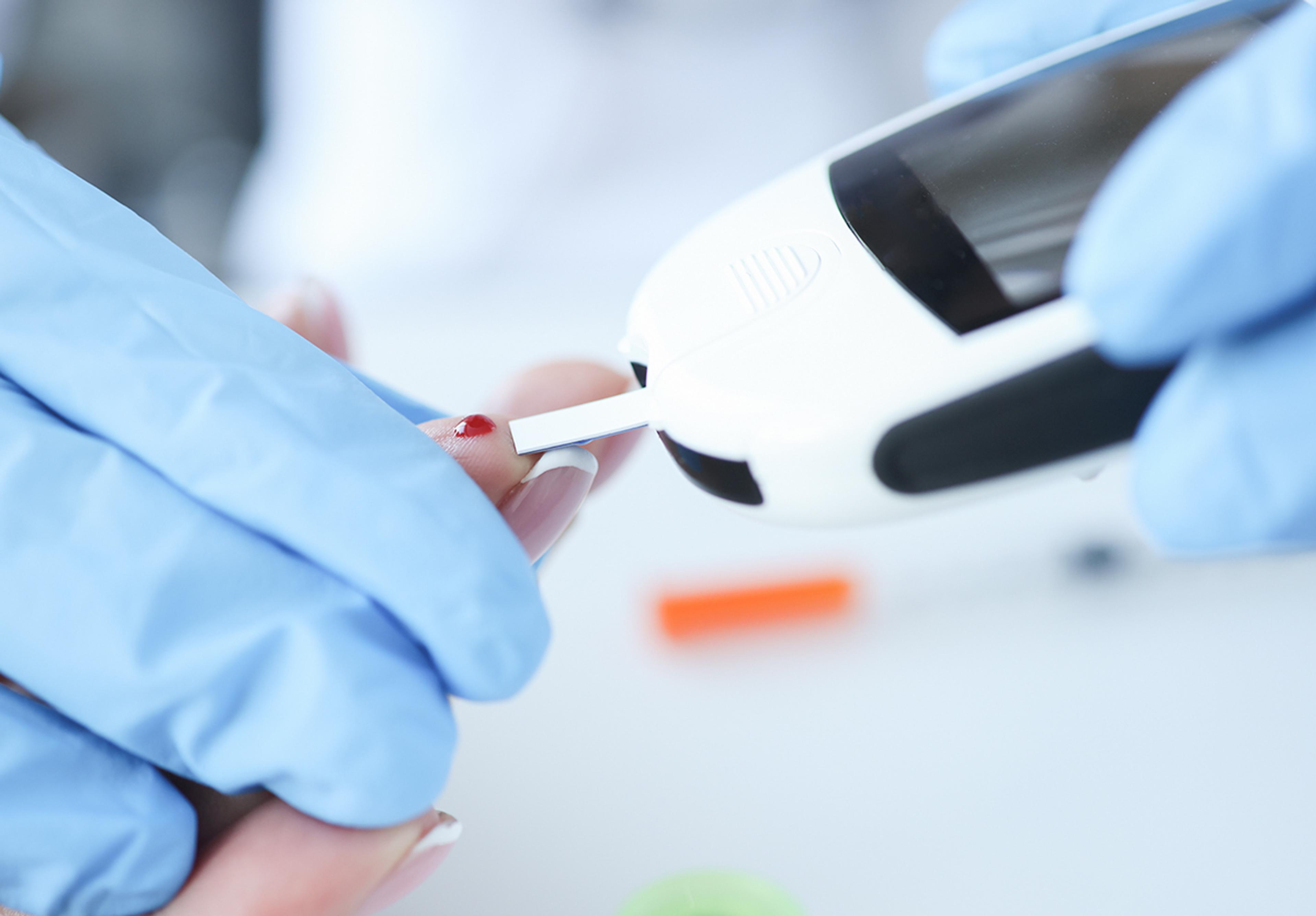 Close-up of a gloved healthcare professional performing a finger-prick blood test, placing a small drop of blood onto a diagnostic test strip using a handheld glucose meter in a clinical or point-of-care setting.