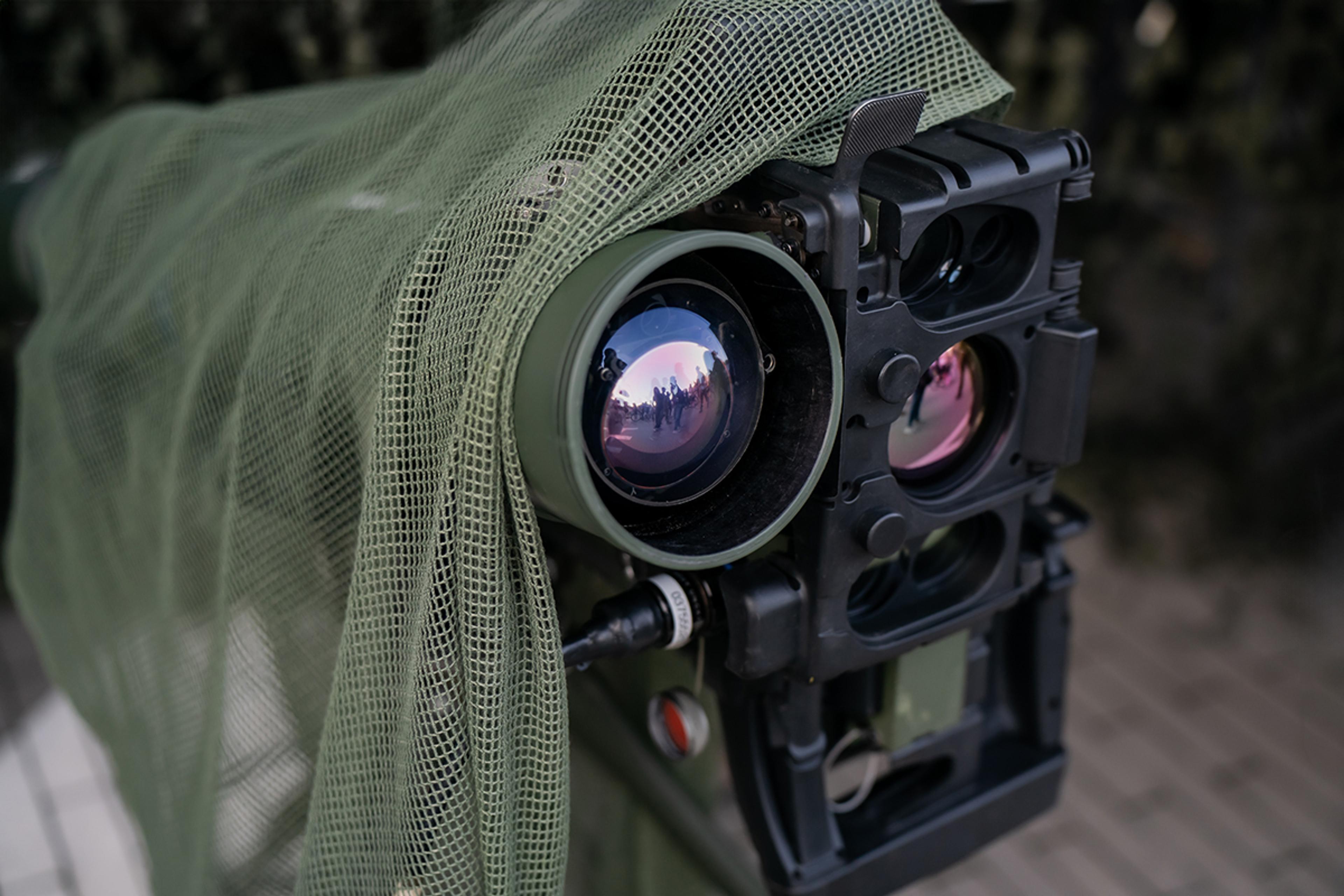 Close-up of a military optical surveillance or targeting system partially covered with green camouflage netting, showing multiple large precision lenses and sensor apertures in a rugged black housing. The image highlights a concealed defence electro-optic payload designed for observation, detection, targeting, or battlefield situational awareness in tactical environments.