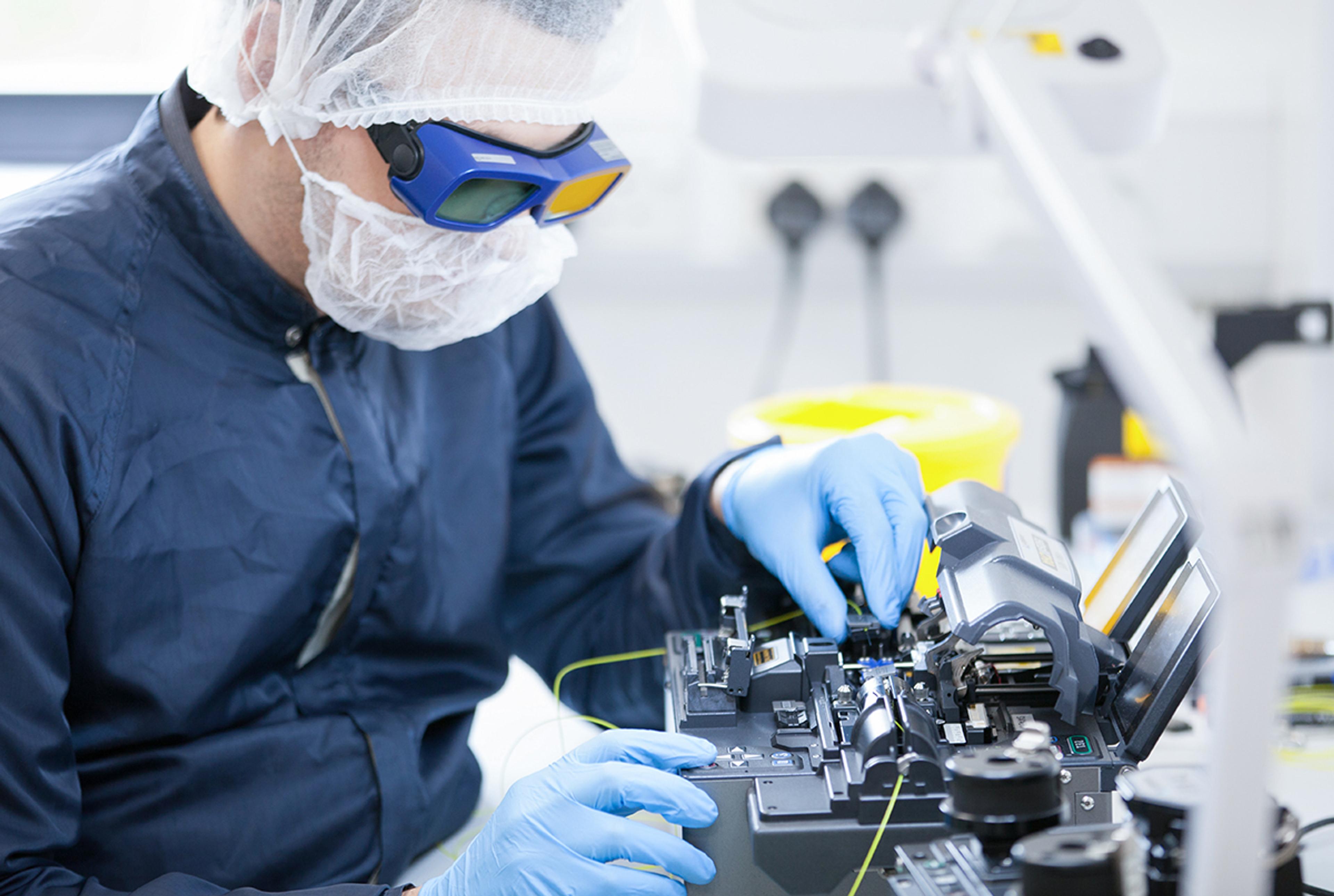 Technician wearing protective eyewear, gloves and cleanroom clothing operates precision fiber optic splicing equipment at the G&H Torquay manufacturing facility.