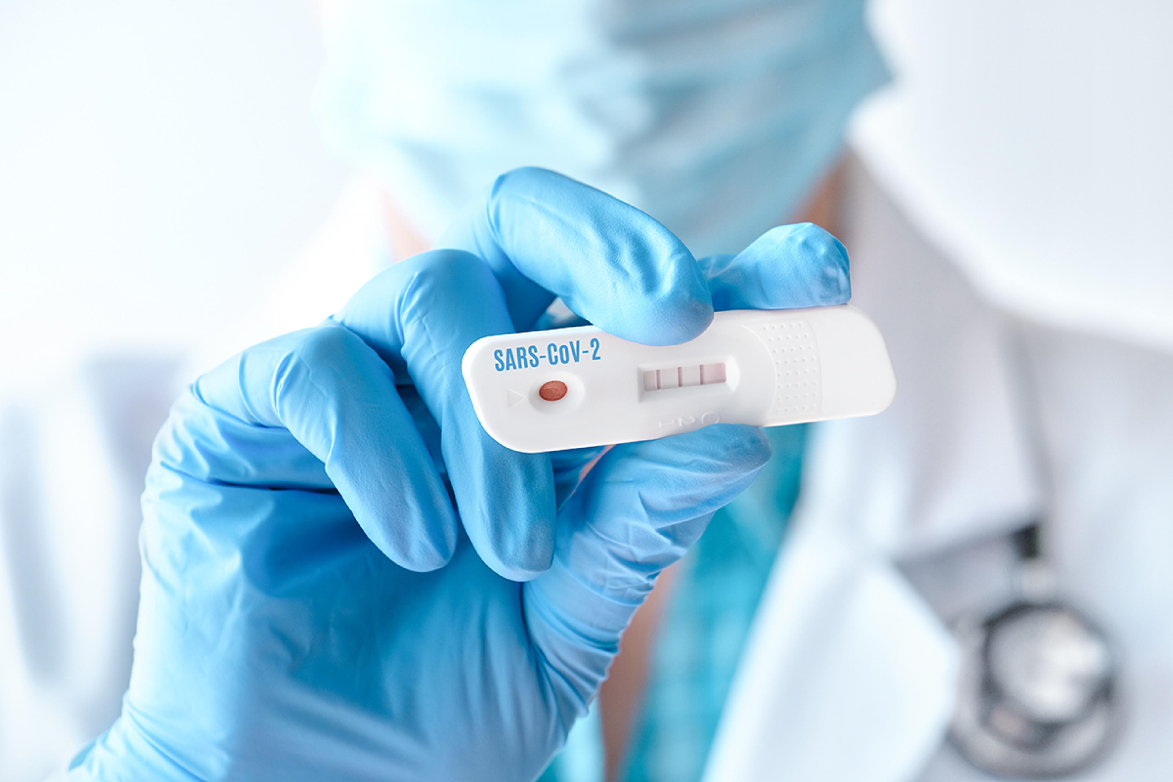 Gloved hand holding a SARS-CoV-2 rapid test cassette showing a positive result, with a clinician blurred in the background.