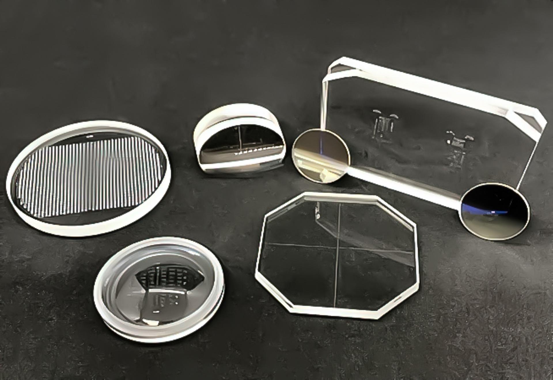 A selection of optical reticles of various shapes and designs arranged on a dark surface. The set includes circular, rectangular, and octagonal glass components with etched patterns, mirrored surfaces, and transparent sections, used in precision optical applications.