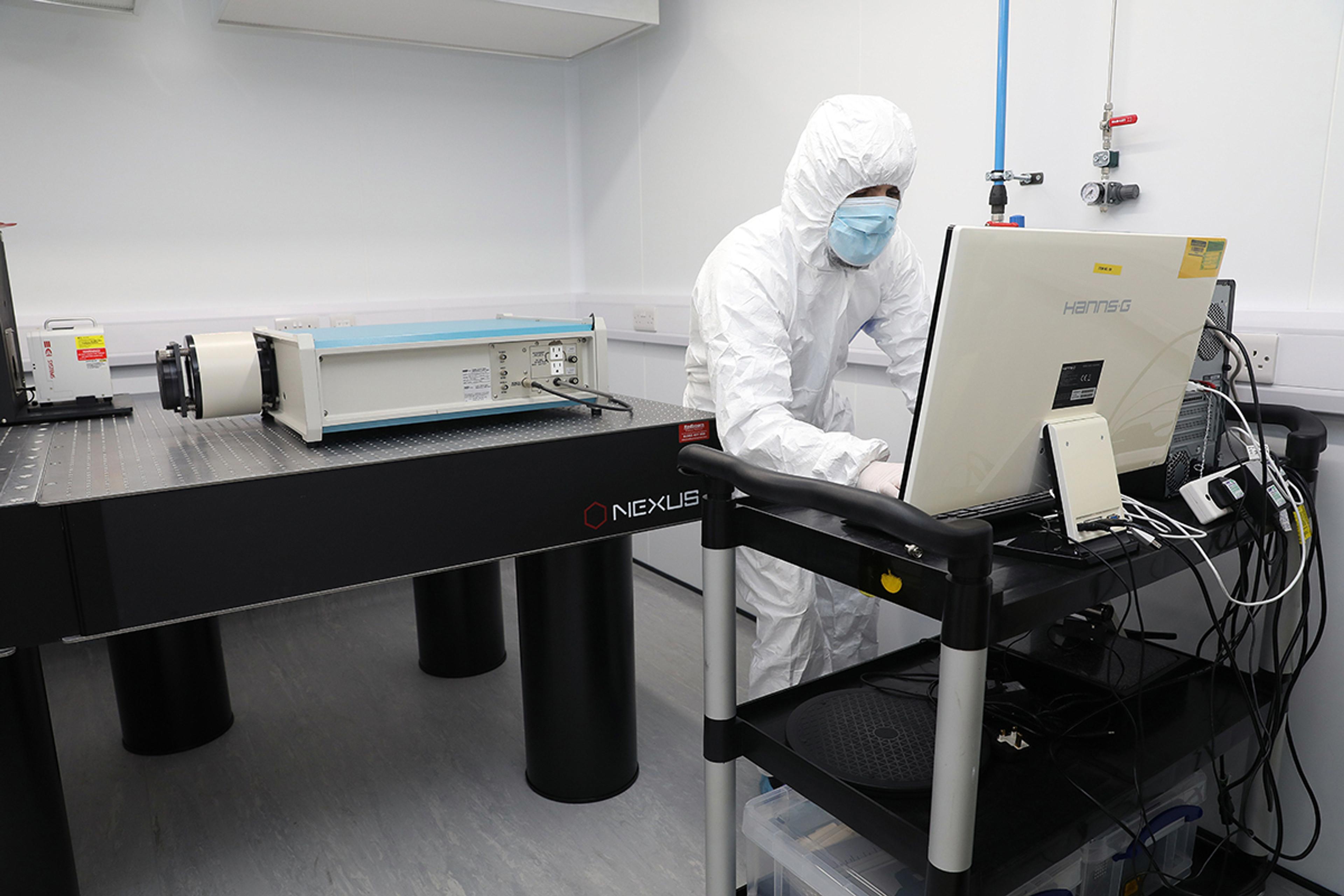 Technician in full cleanroom suit and mask operating a computer workstation beside a large optical instrument on an anti-vibration Nexus lab table in a sterile laboratory setting.