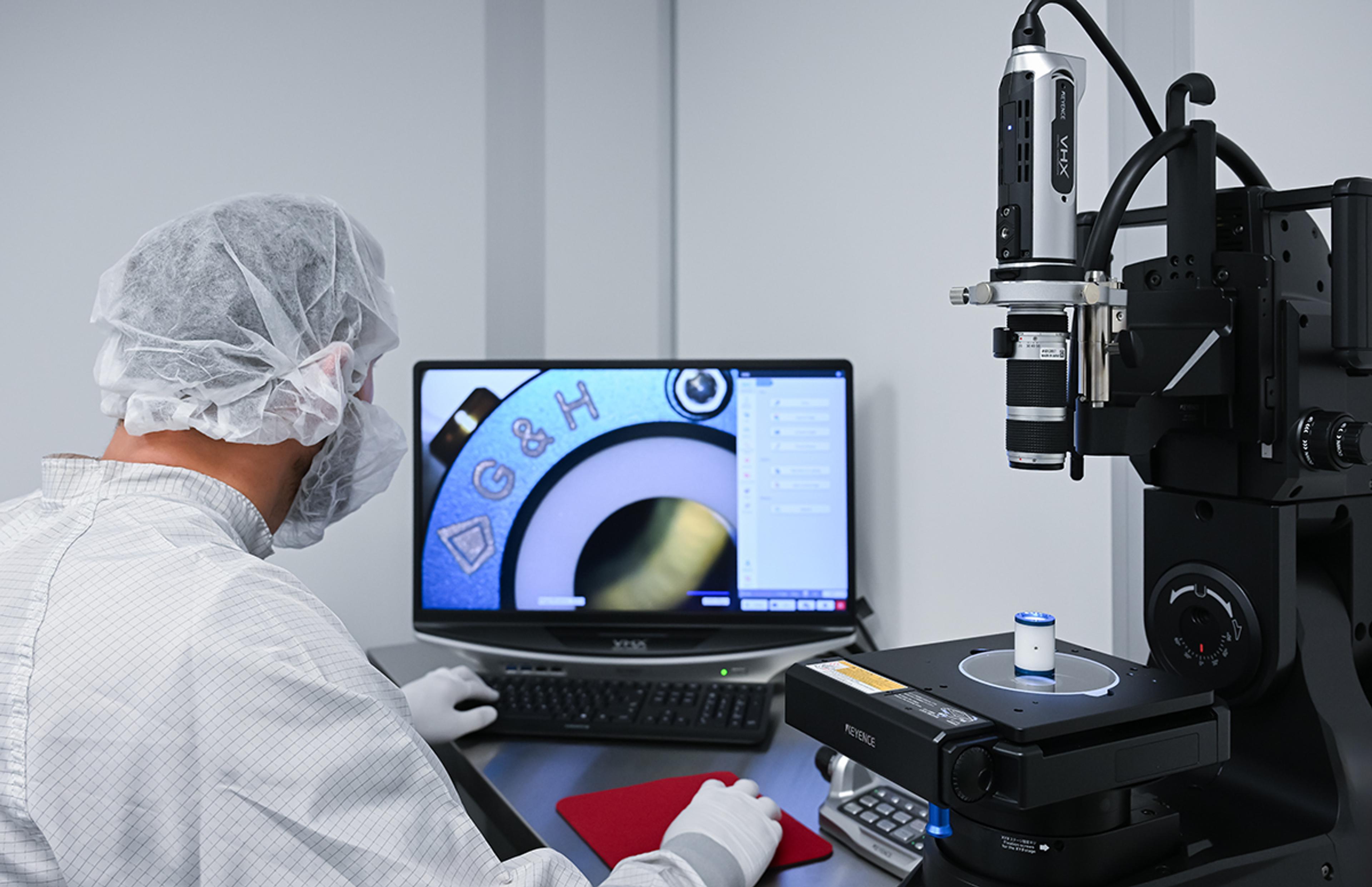 Technician in cleanroom attire operates an optical inspection system, viewing a magnified image of a G&H-branded pockels cell precision component on a computer monitor, with a Keyence digital microscope in the foreground.