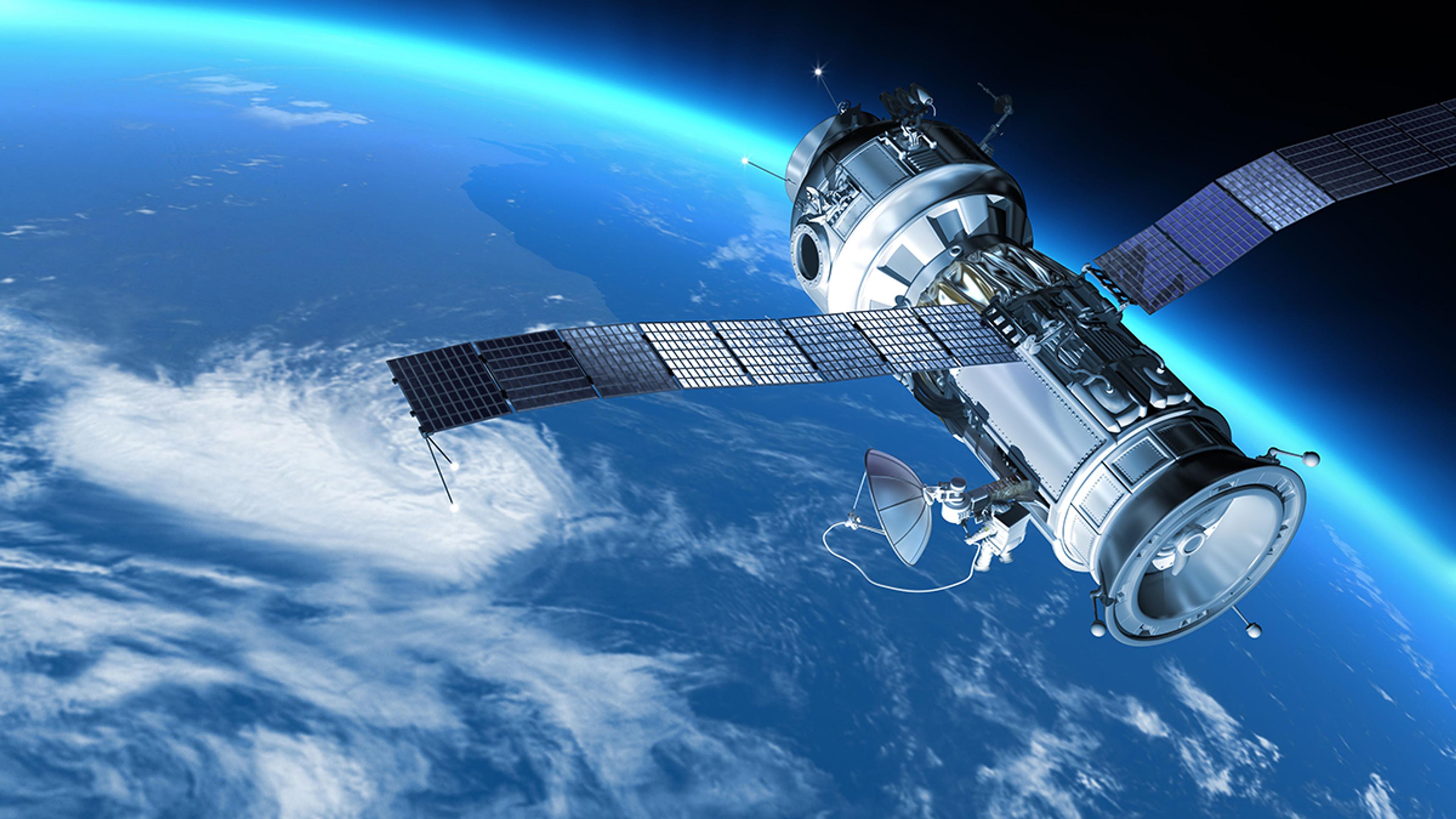 A satellite orbiting Earth with large solar panel wings extended on either side. The satellite has a cylindrical body with various antennas and sensors, including a dish antenna. Below, the Earth's curved horizon is visible with a vivid blue atmospheric glow, white clouds, and landmasses partially covered by ocean. The image conveys a sense of high-altitude space observation and advanced technology.