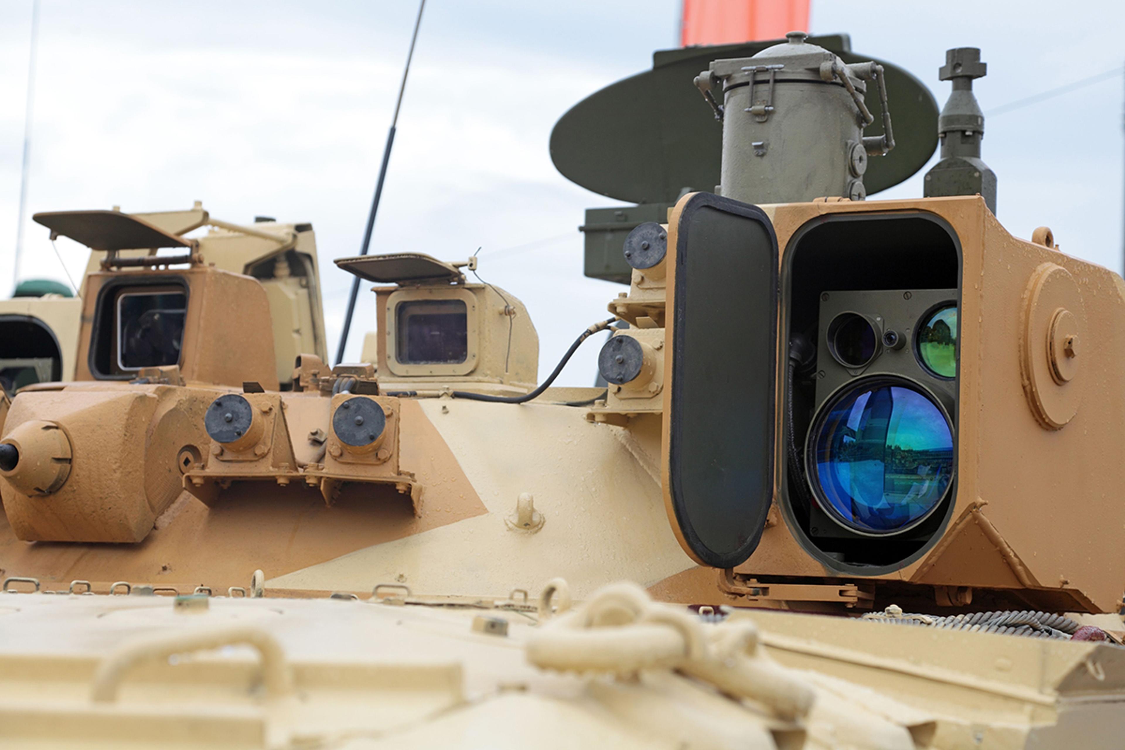 Close-up of a military armored vehicle equipped with advanced optical and sensor systems. The image highlights a large lens and multiple viewing apertures mounted on the tan-colored exterior, likely used for targeting or surveillance. The setting appears to be an outdoor exhibition or display area.
