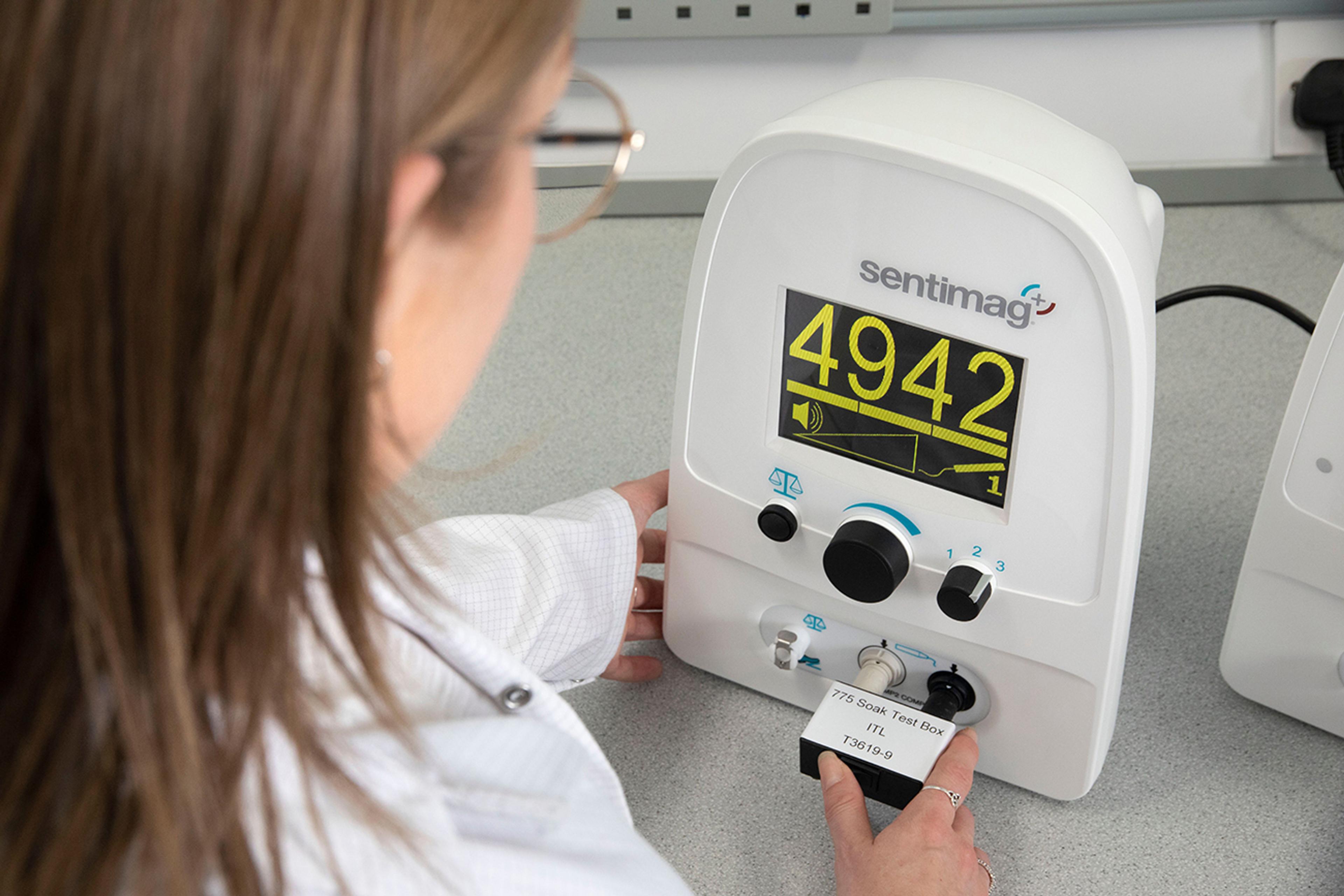Testing a medical device at ITL Medical - A person wearing a lab coat is operating a Sentimag medical device with a large digital display showing the number 4942. The user is inserting or adjusting a labeled test component at the bottom of the device, suggesting a calibration or diagnostic procedure in a clinical or laboratory setting.