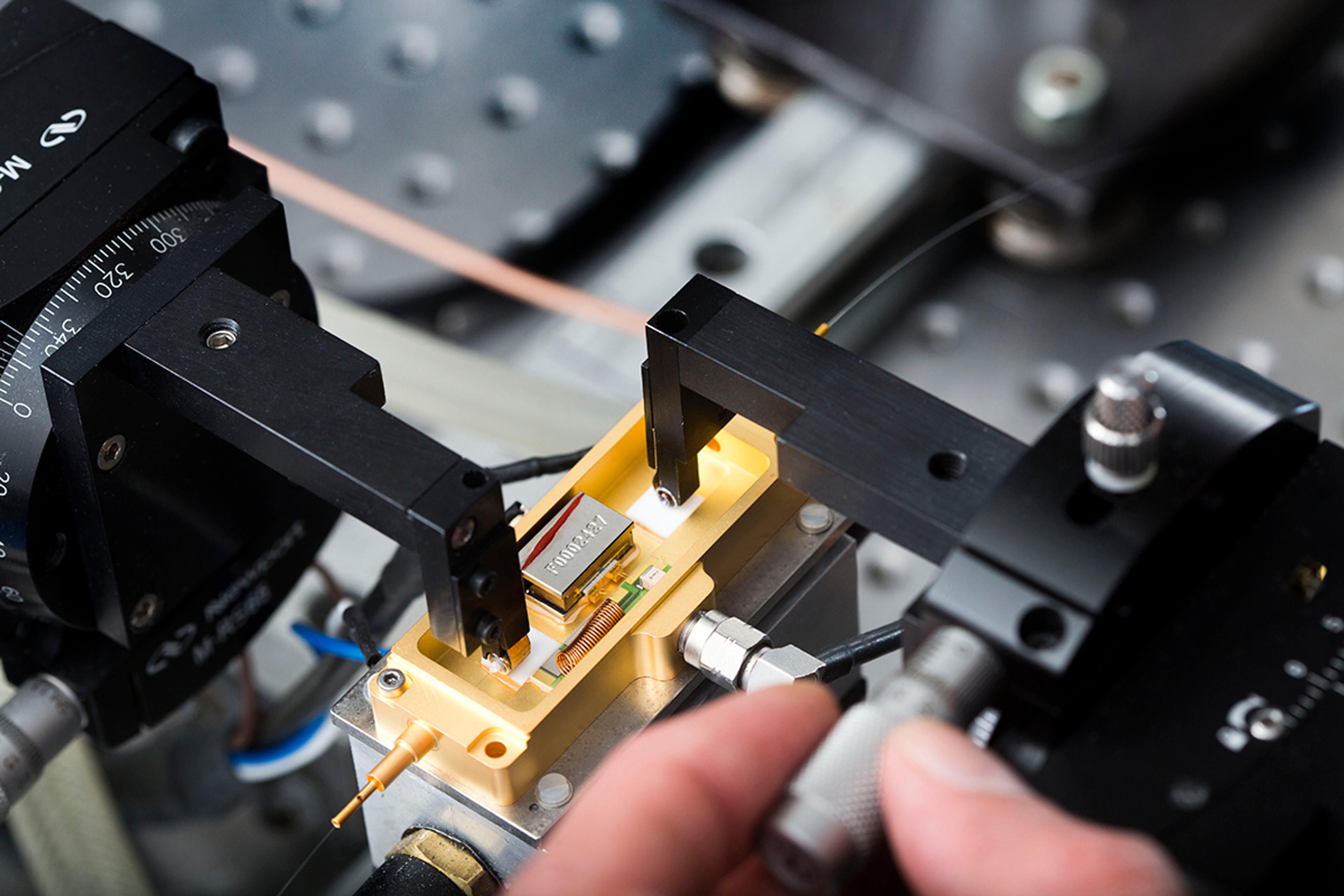 Precision test setup measuring an open gold fibre-coupled laser module on a fixture, with probe arms and a technician adjusting a micrometer stage.