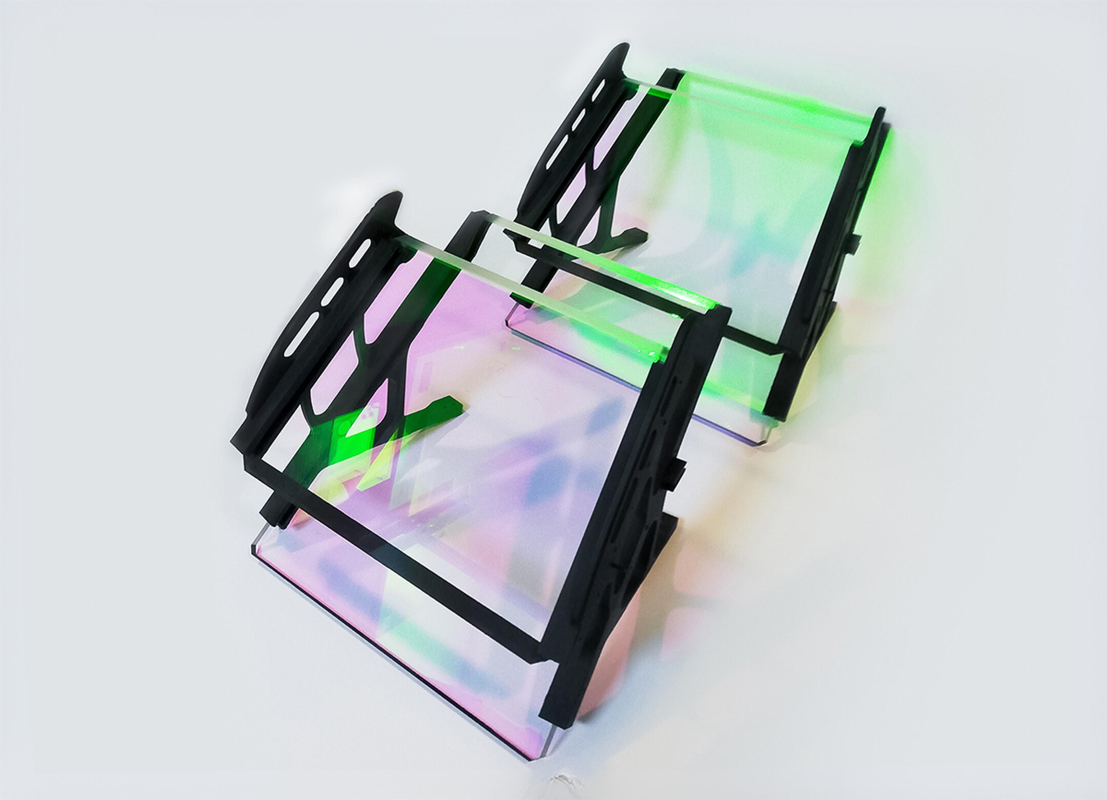 Helmet mounted display system (HMD), Two optical display assemblies featuring precision-coated transparent glass panels mounted in black structural frames. The glass surfaces reflect vivid green, pink, and purple hues, indicative of specialized optical coatings. The assemblies are placed on a light background, and their reflections create colorful patterns beneath them. These components are likely used in augmented reality (AR) or head-up display (HUD) systems, where precise light manipulation is critical