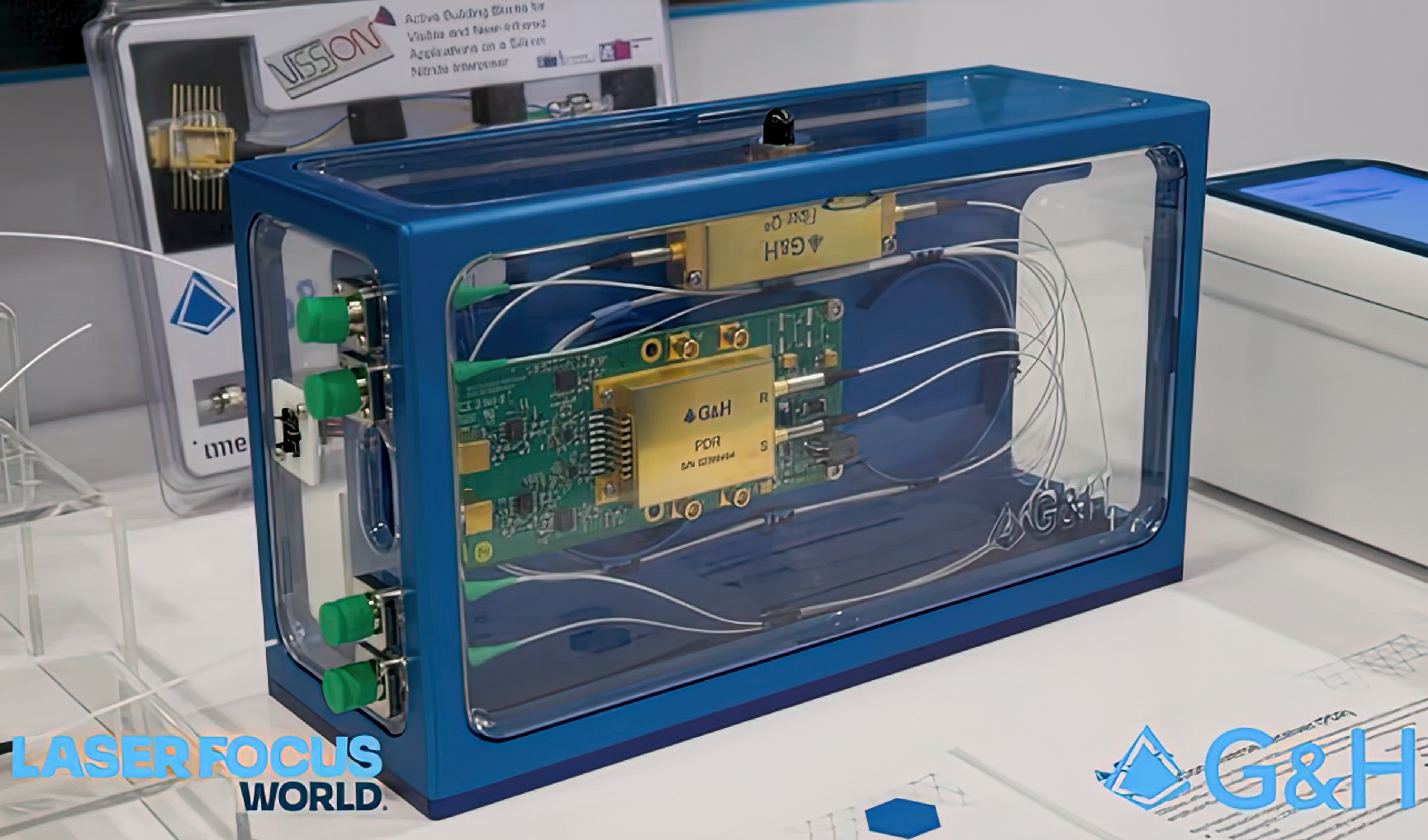Close-up of a G&H photonics assembly inside a clear blue display case, showing fibre-connected modules and circuit boards, with Laser Focus World and G&H logos visible.