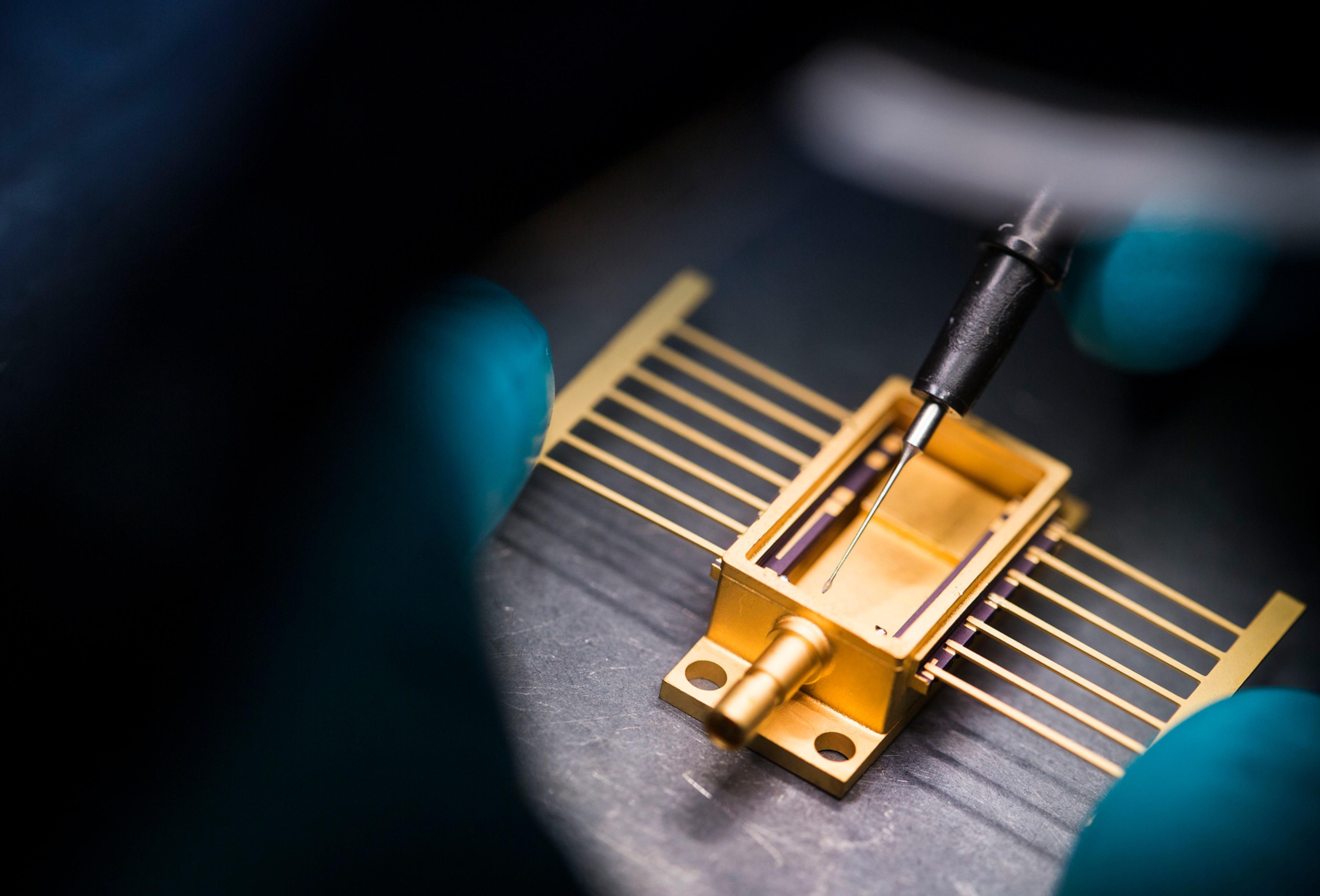 A close-up image of a precision electronic component being assembled or inspected. The gold-colored device has multiple fine wire leads extending from its sides, and a probe or fine tool is positioned above it. Gloved hands are visible, indicating a cleanroom or high-tech laboratory environment
