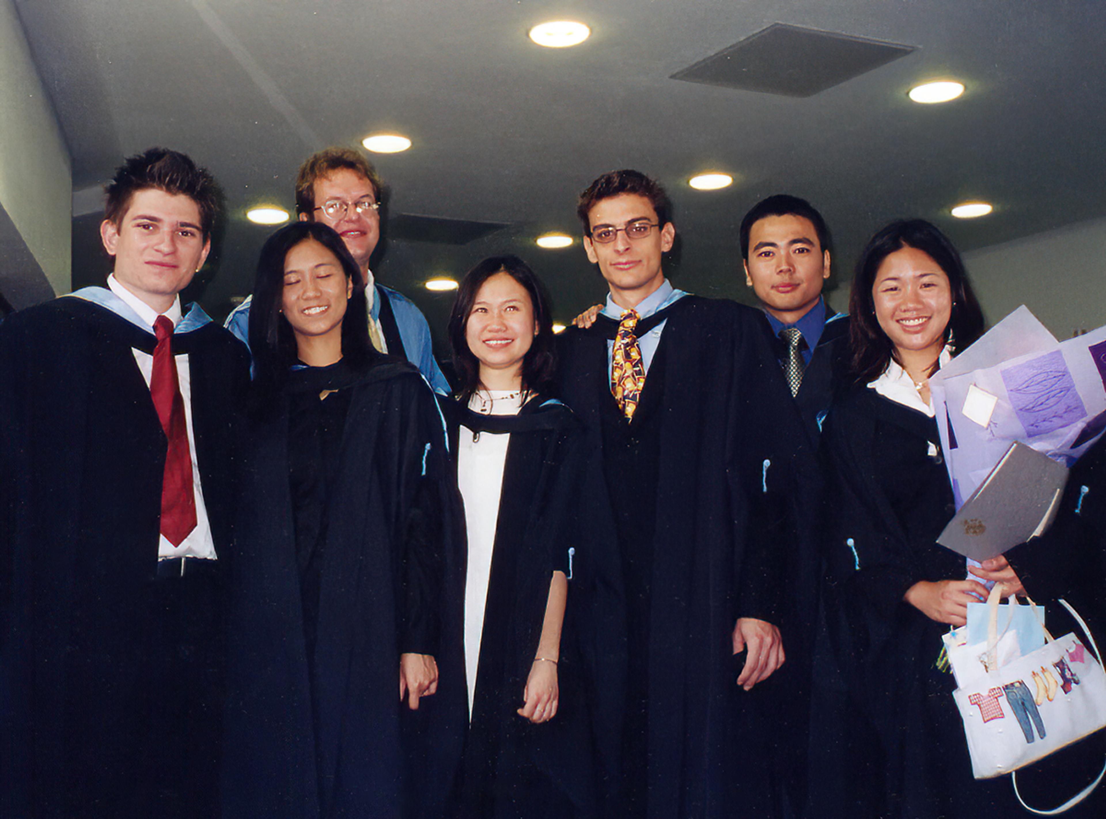 Seven people posing together indoors at a graduation celebration, wearing academic gowns, with one holding flowers and a certificate folder.