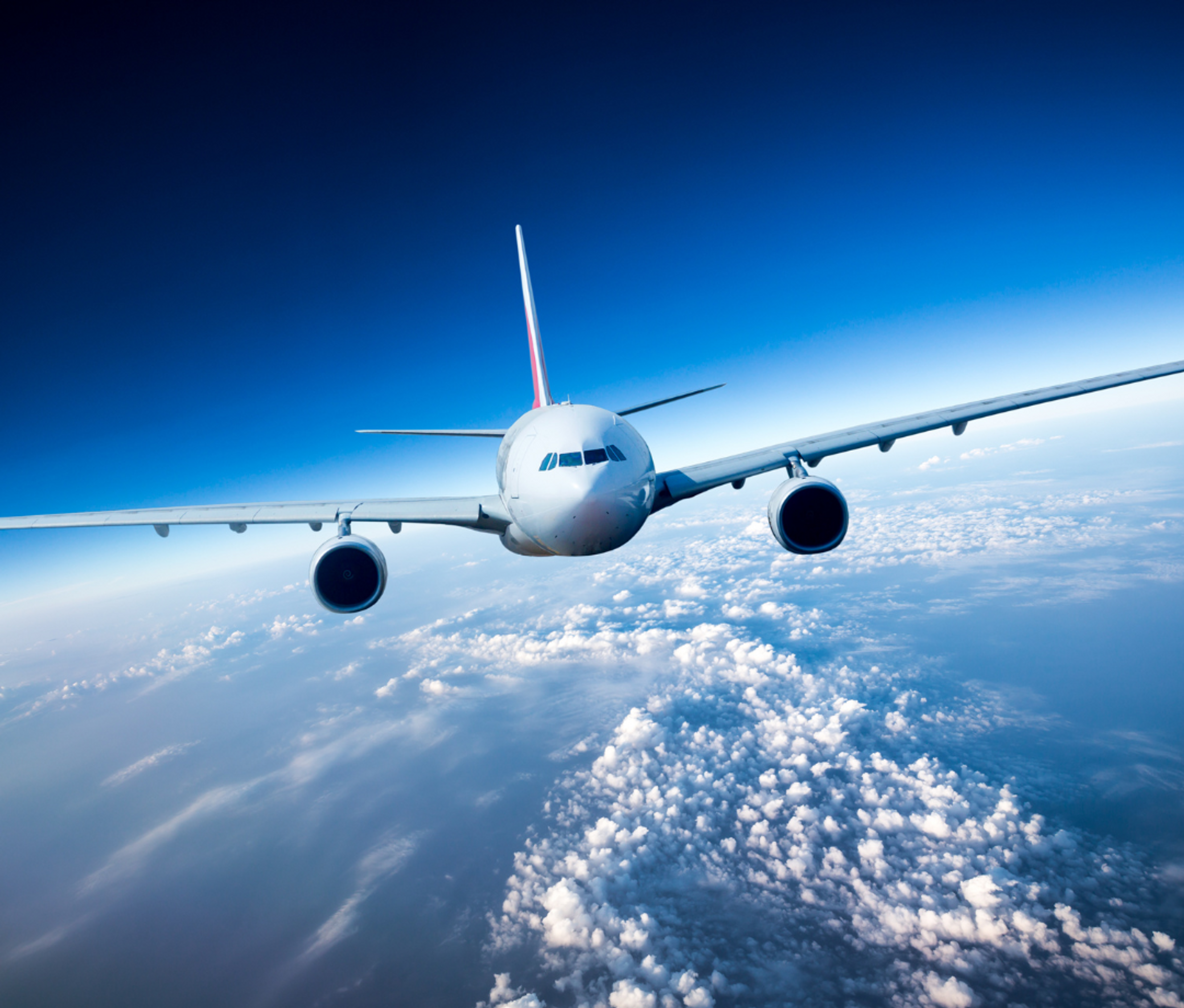 Front view of a commercial Boeing aircraft flying above the clouds with a vivid blue sky background, captured mid-flight from a slightly lower angle.