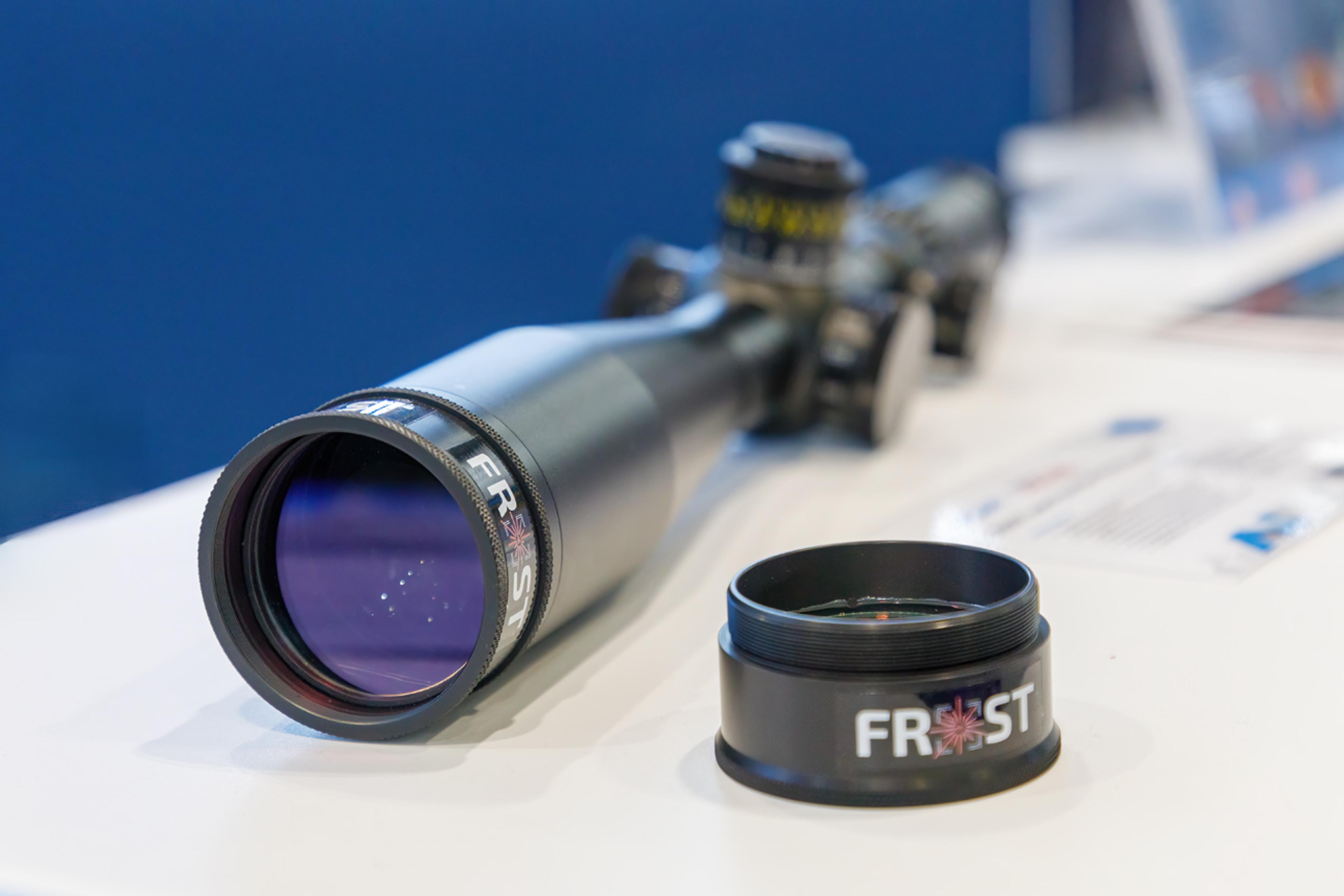 Close-up of a riflescope fitted with a G&H FROST laser protection filter, designed to safeguard optical systems from laser threats. The black cylindrical filter with the FROST logo is shown attached to the front of the scope, with an additional filter cap placed nearby on a white surface.