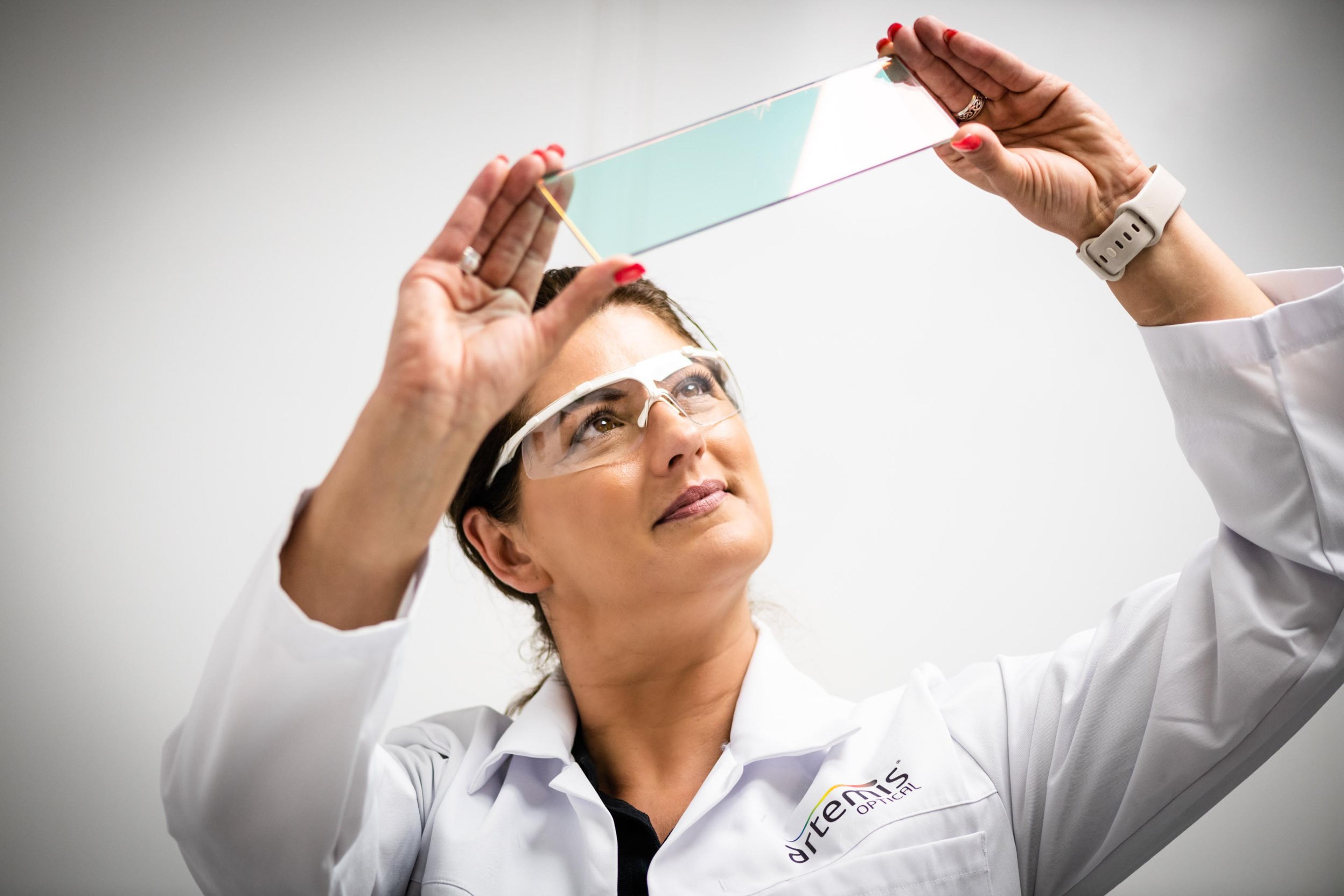 A female technician wearing safety glasses and a white lab coat with the Artemis Optical logo inspects a large, rectangular optical filter with an iridescent coating, holding it up to the light in a clean, bright laboratory environment.