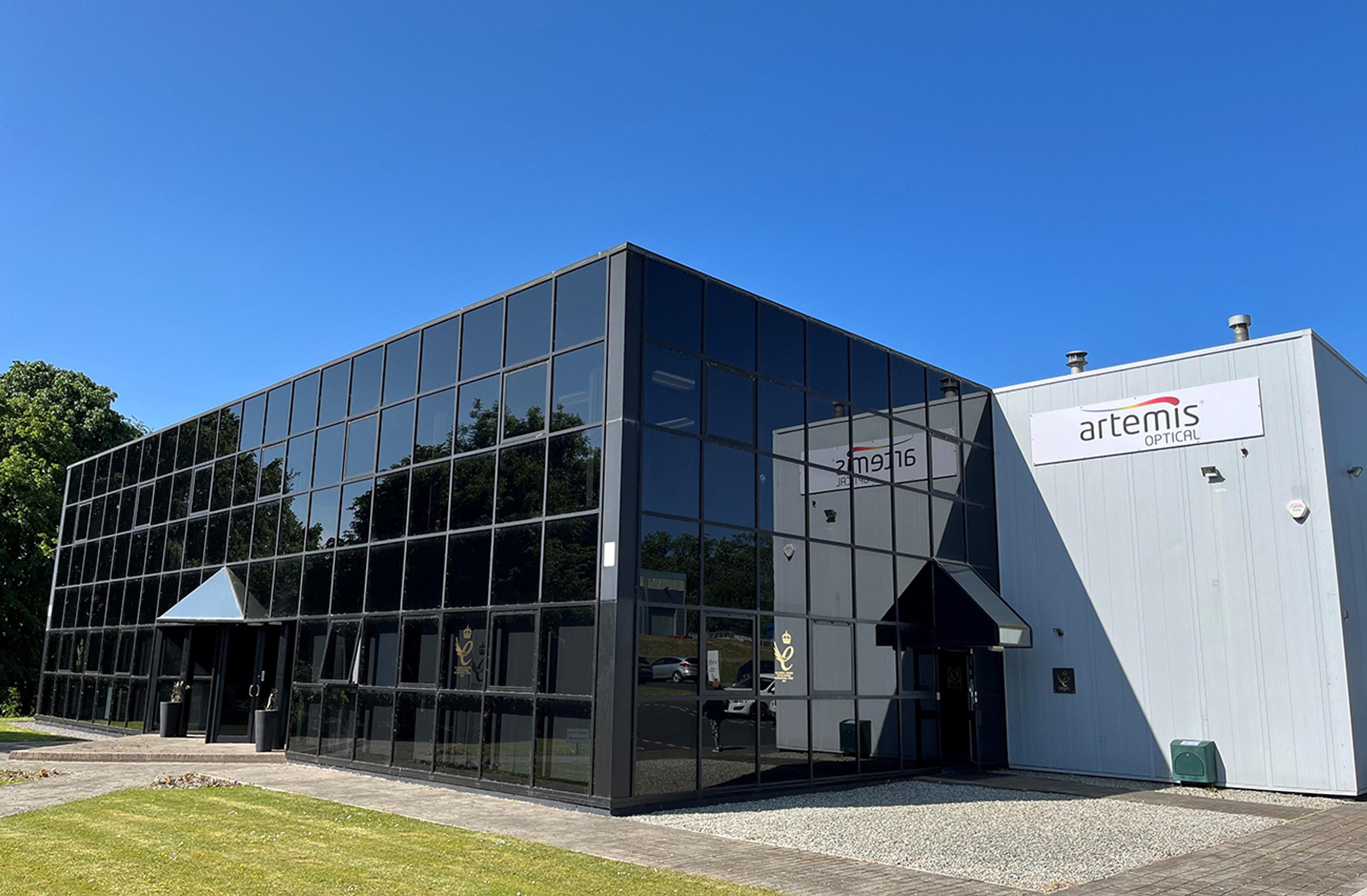 G&H | Artemis in Plymouth, UK - Modern industrial building with reflective glass façade and a white extension featuring the 'Artemis Optical' logo. The structure is surrounded by a well-kept lawn, under a clear blue sky