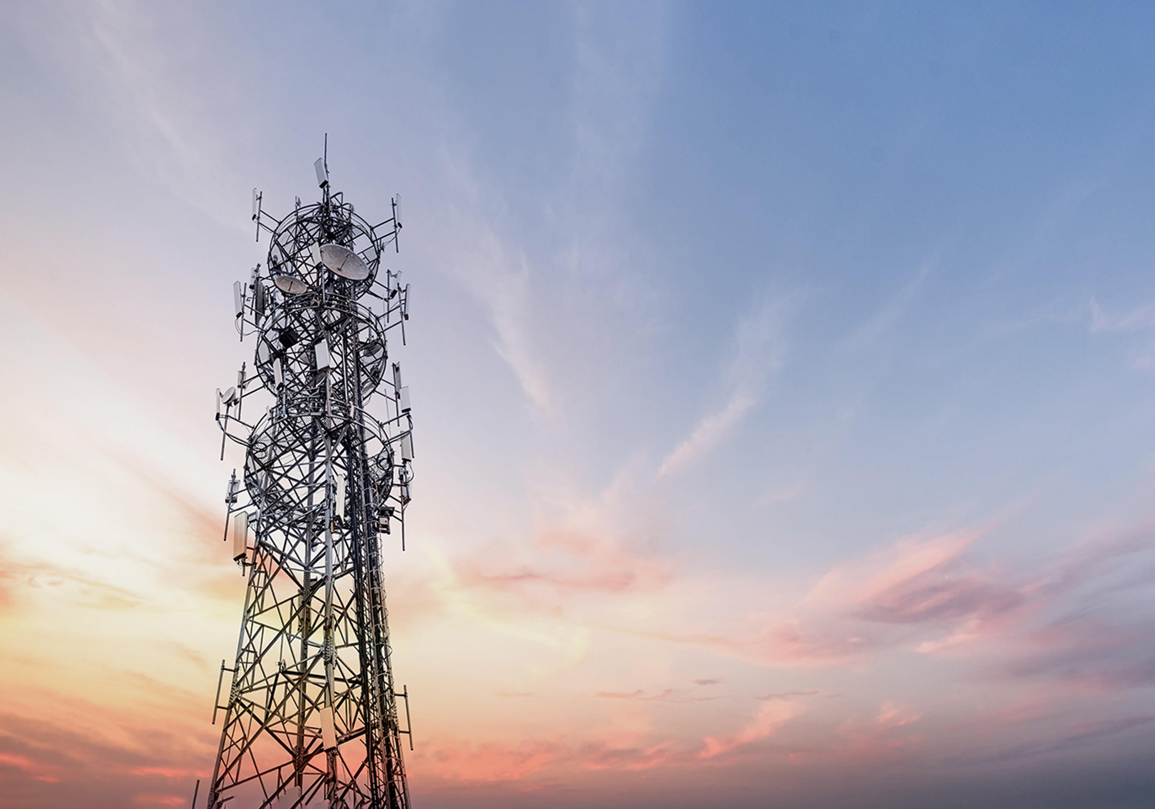 Telecommunications tower with multiple antennas and dishes silhouetted against a colourful sunset sky.