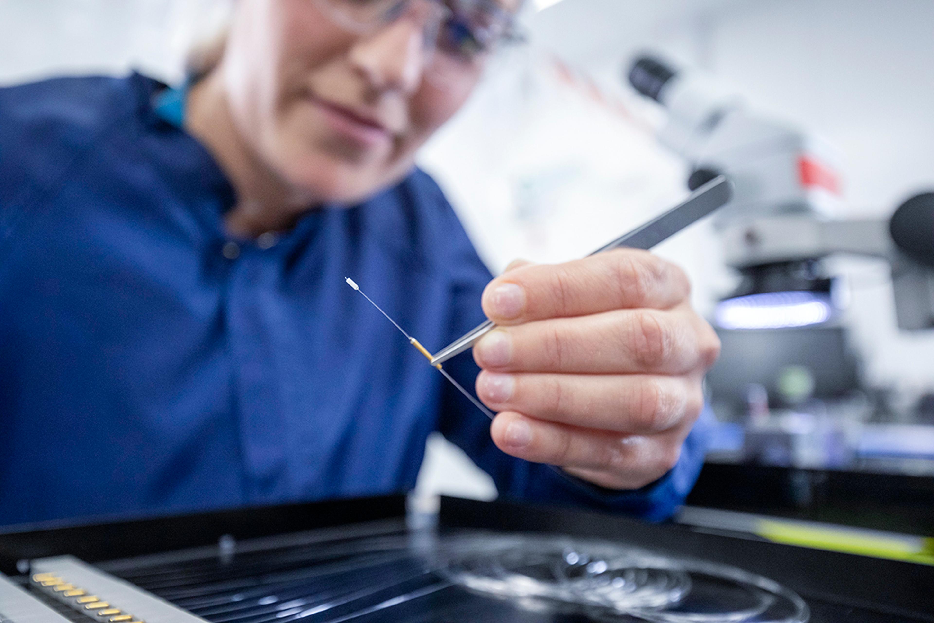 Technician uses precision tweezers to inspect and handle a fine fiber optic component beside a microscope at the G&H Torquay facility.