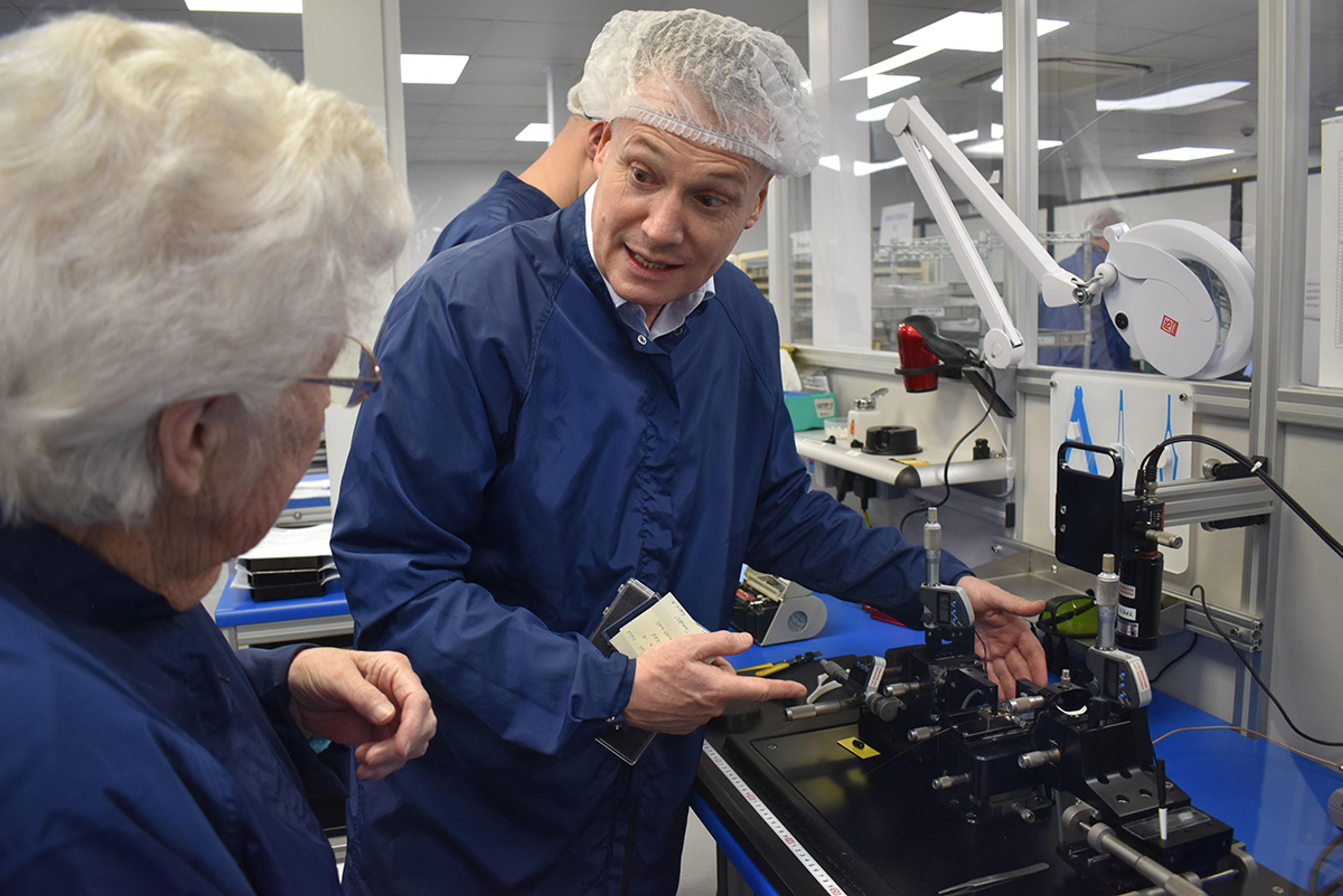 Norma Knapp & Toby Reid, Product Management Director, G&H Torquay. Inside a cleanroom lab, a man wearing a blue lab coat and hairnet enthusiastically explains a technical setup on a black optical bench to an elderly woman also in a lab coat. The table is filled with precision instruments and measurement devices, likely used for optical alignment or testing. The environment is brightly lit, with additional lab equipment and organized workstations visible in the background. The interaction appears to be part of a tour or demonstration.