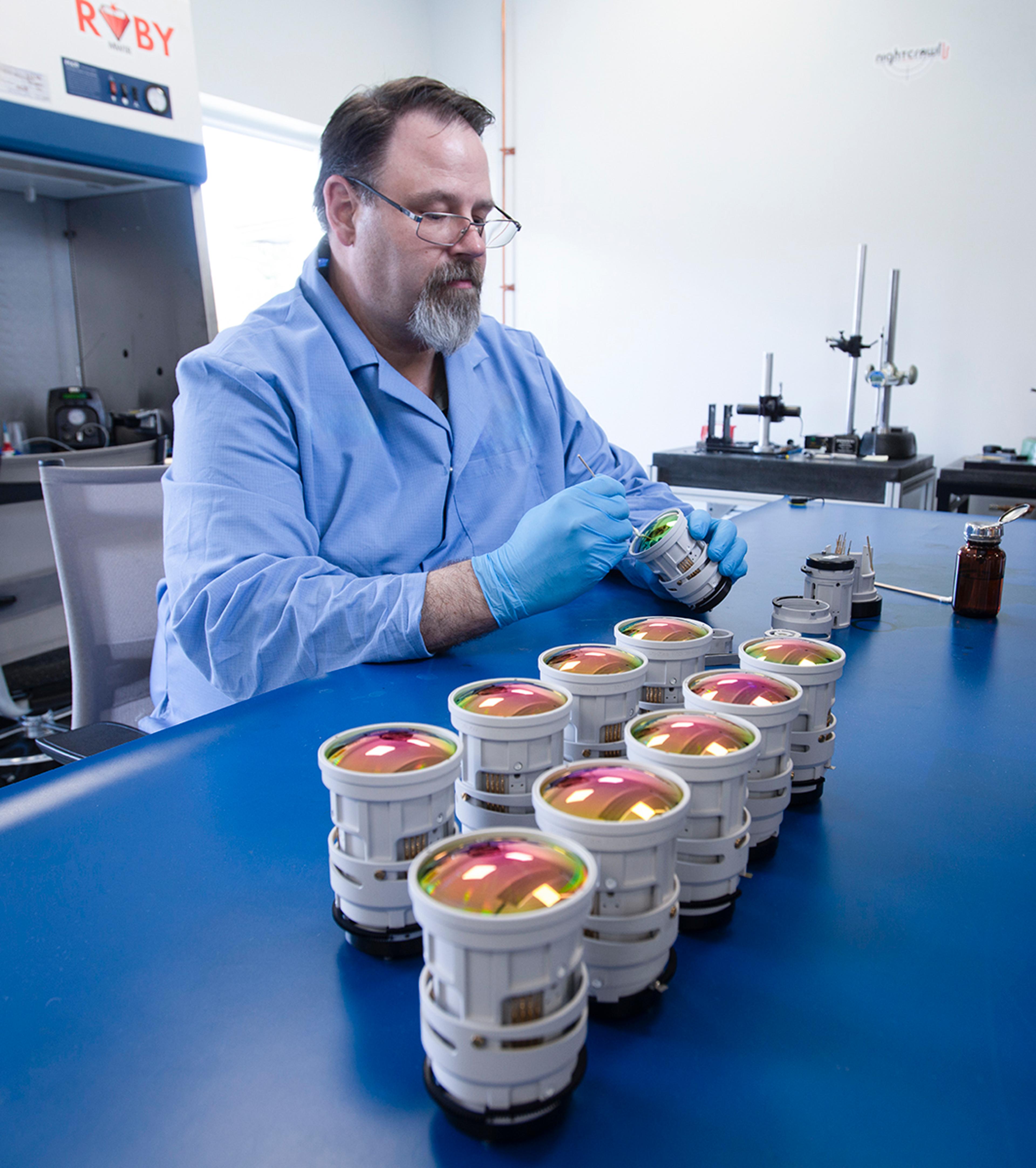 G&H technician in protective clothing and gloves assembling precision Stingray lens systems, with multiple coated optical lens assemblies arranged on the workbench.