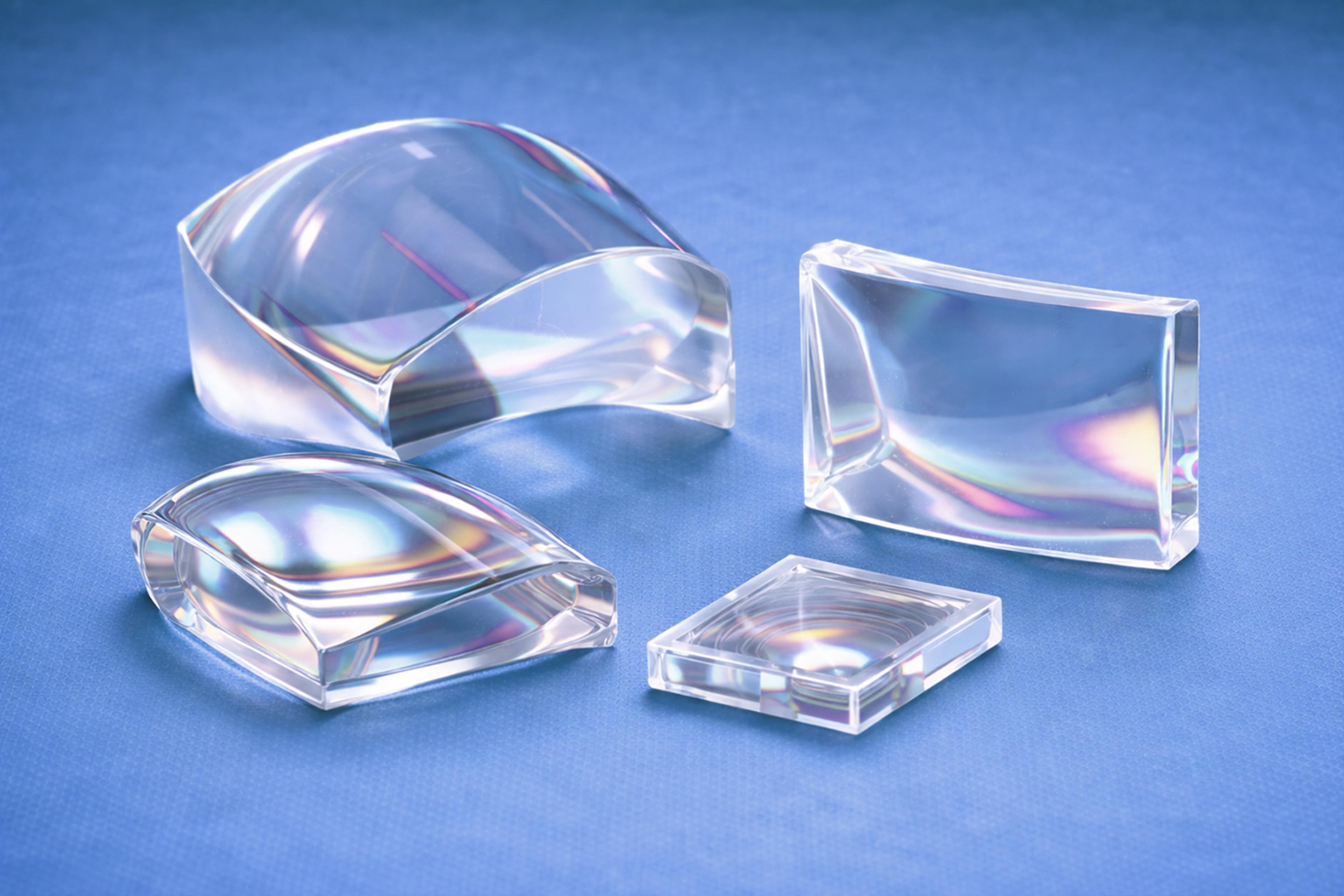 Selection of transparent freeform polymer optical components with curved and asymmetric surfaces, displayed on a blue background to illustrate advanced freeform optics for precision light shaping and imaging applications.