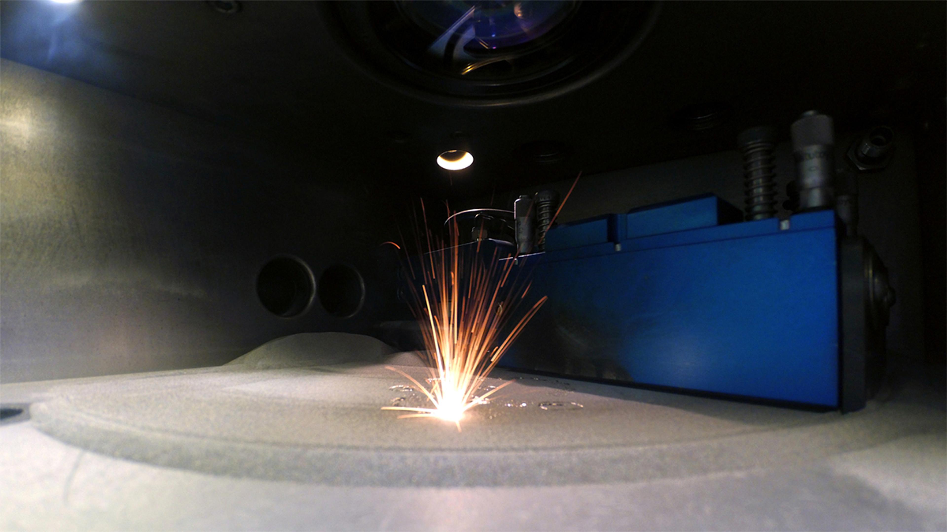 Close-up view inside a metal 3D printing or laser sintering machine during operation. Bright sparks fly upward as a focused laser fuses metal powder on a build platform. The environment is industrial and enclosed, with metallic surfaces, circular vents, and a prominent blue component housing. The scene highlights advanced additive manufacturing technology used in precision engineering and prototyping