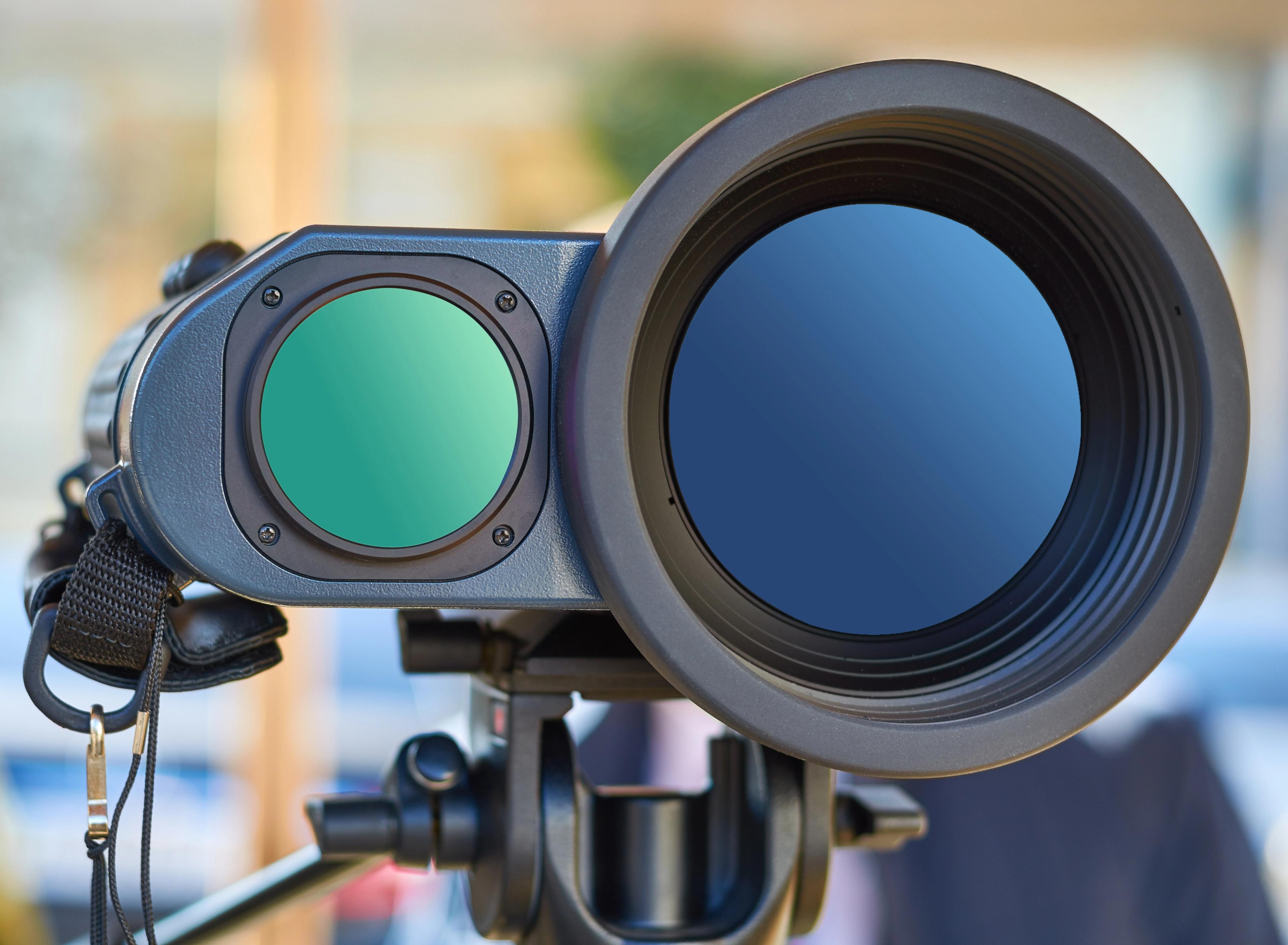 Close-up front view of a tripod-mounted optical surveillance or targeting device with a large main lens and a smaller green-tinted lens beside it.
