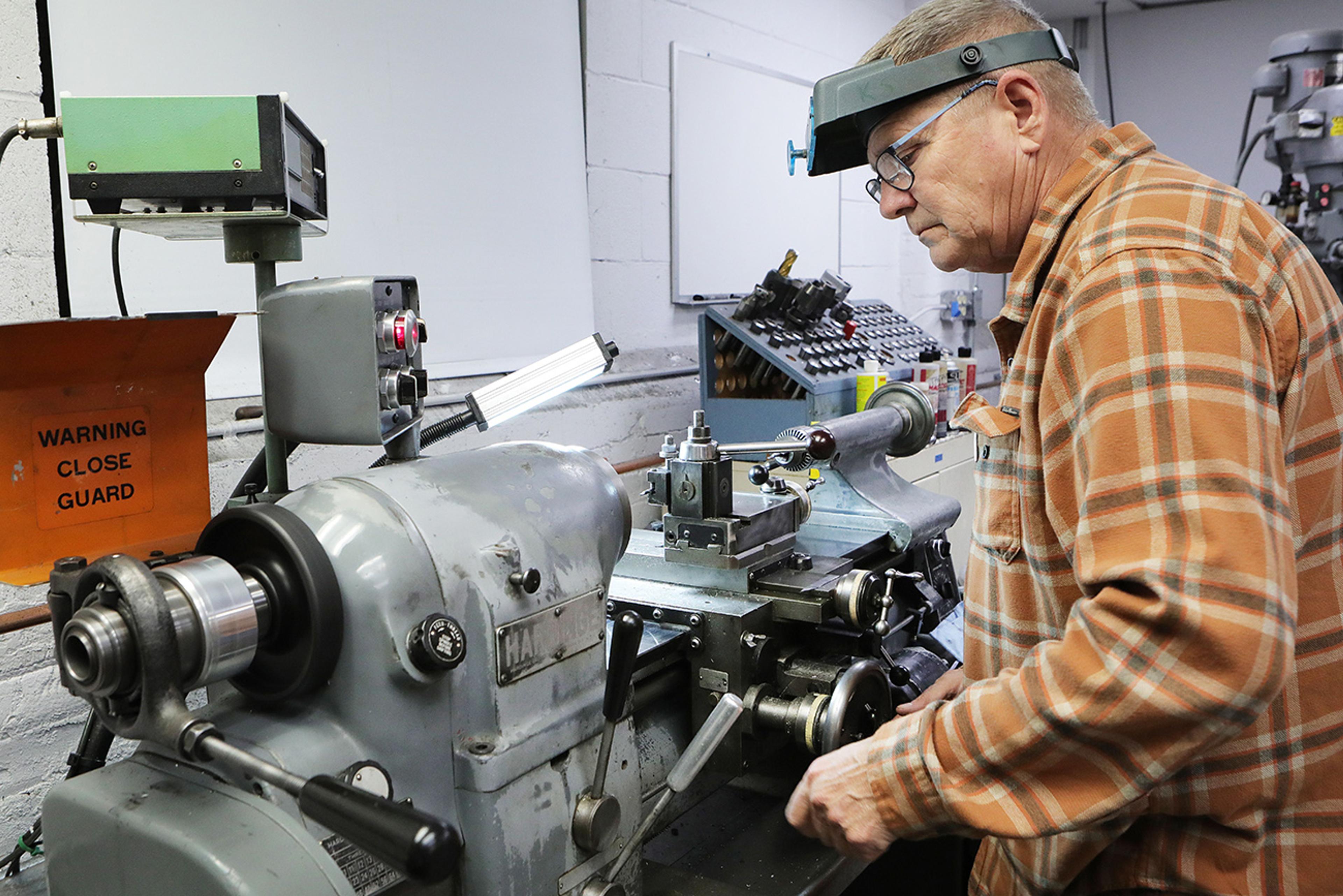 Manufacturing of polymer optics, An older man wearing a plaid shirt and magnifying headband operates a metal lathe in a workshop. The machinery is surrounded by tools and equipment, and a safety sign reading "WARNING CLOSE GUARD" is visible. The man is focused on precision work, suggesting a task requiring detailed manual machining or metalworking