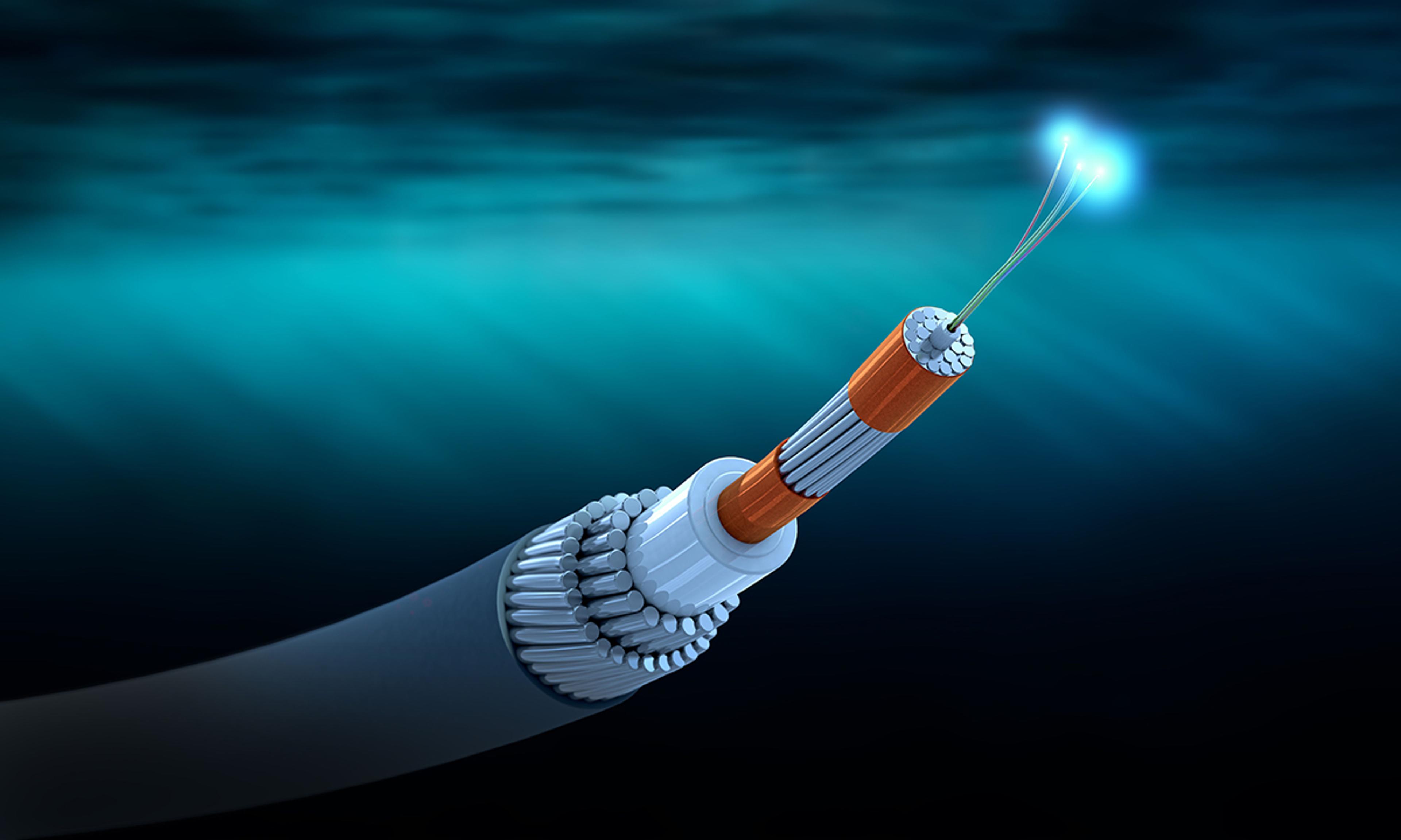 A detailed digital illustration of an undersea fiber optic cable is shown with its layers exposed. The cable is submerged underwater, with light rays filtering from above. The outer layer is black and cylindrical, encasing several internal protective layers. Near the tip, the orange core reveals bundled metallic and insulating layers. Three thin optical fibers extend from the center, emitting glowing blue light, symbolizing data transmission. The background features a deep ocean gradient from turquoise to dark blue.