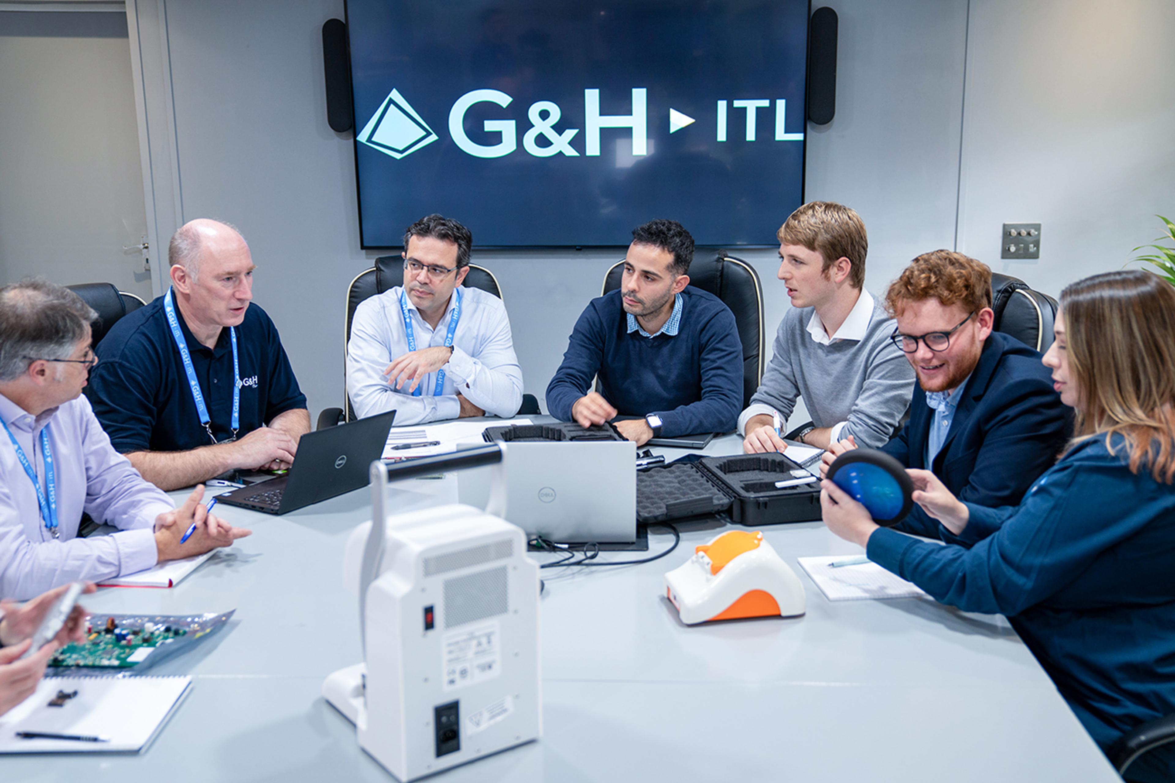 G&H | ITL team in a meeting room collaborating around a table with laptops and prototype devices, beneath a screen displaying the G&H | ITL logo.