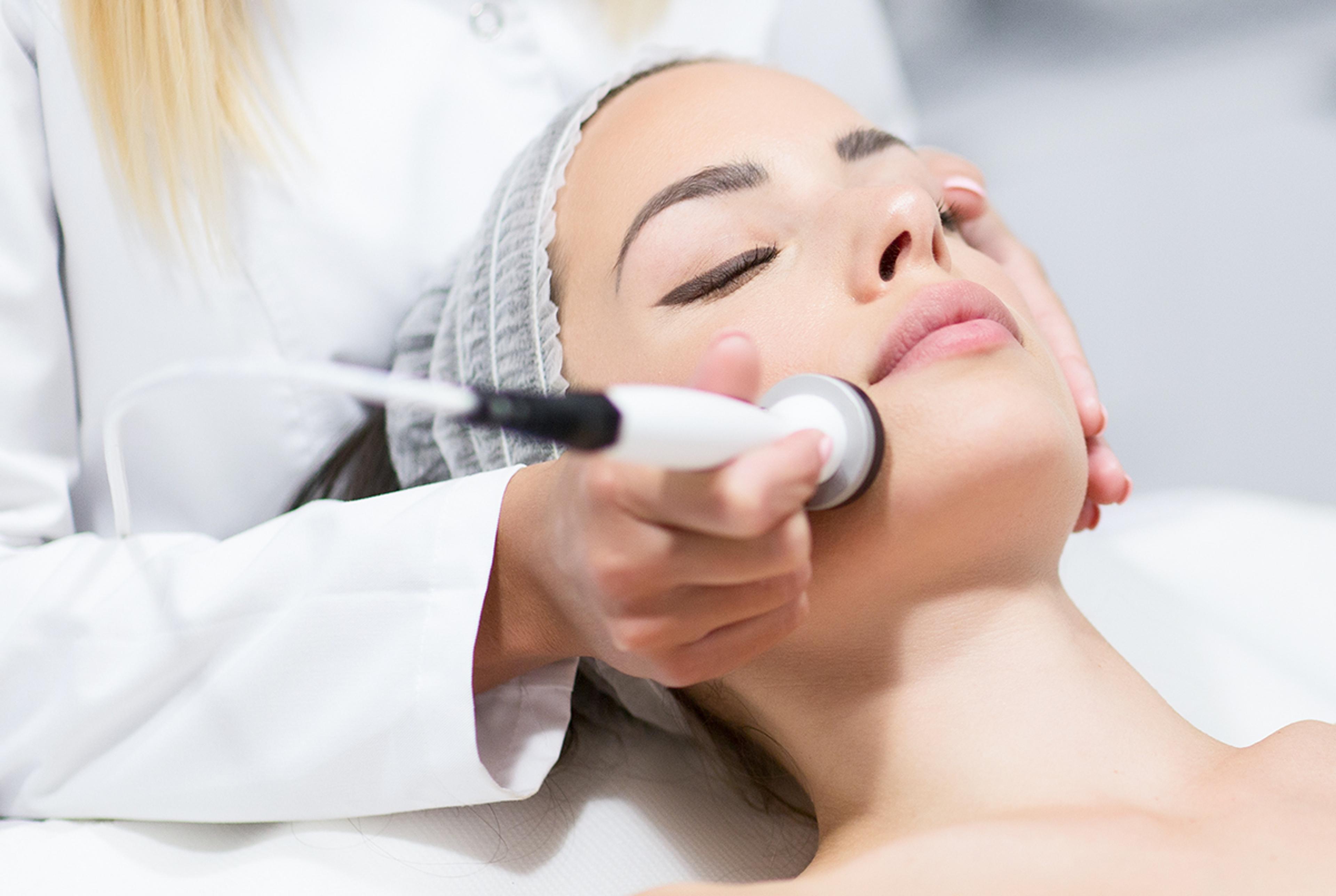 Woman lying back during a facial treatment while a clinician applies a handheld aesthetic device to her cheek.