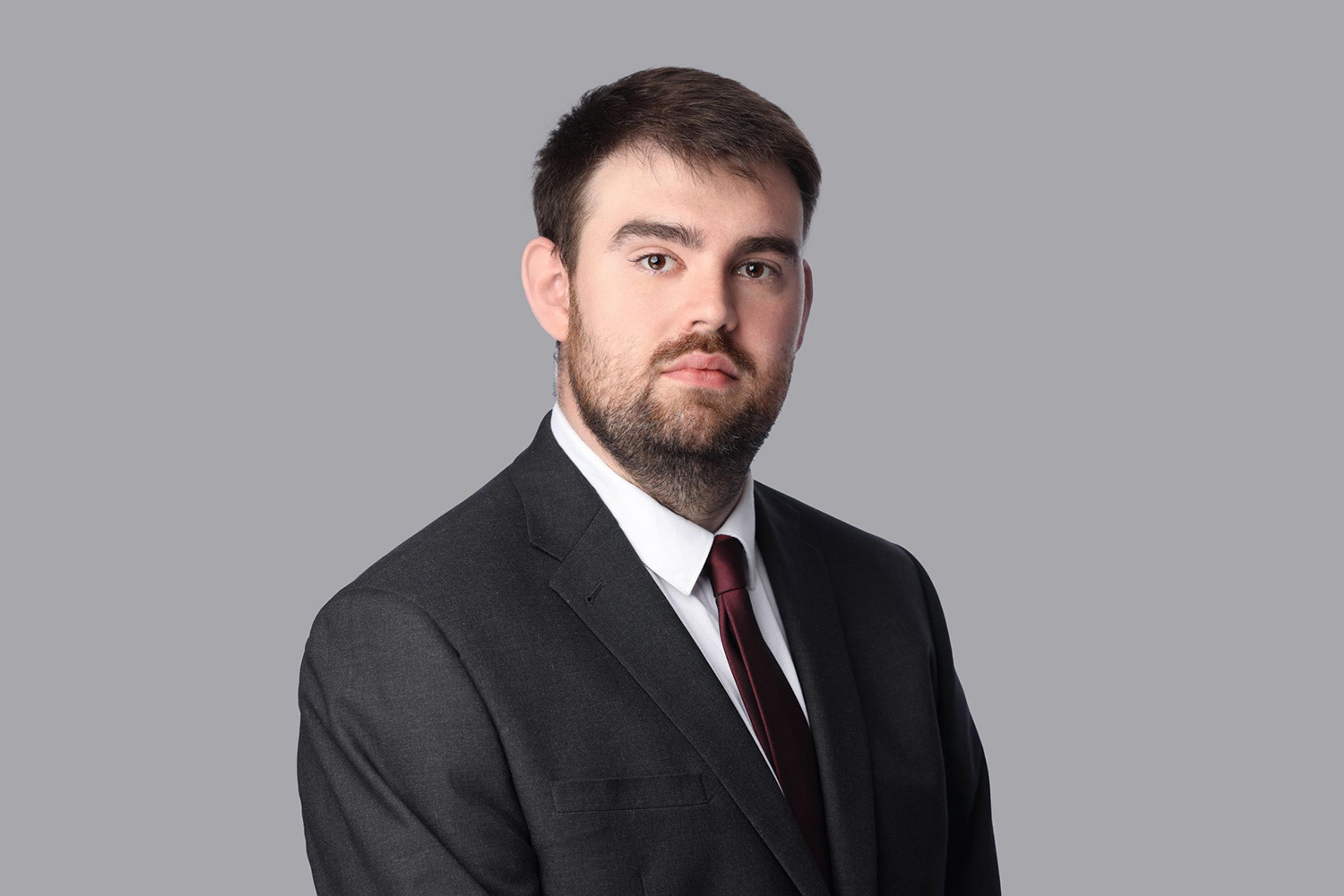 Tom Ackrill - Business Development Manager, A professional headshot of a man wearing a dark grey suit, white dress shirt, and maroon tie. He has short dark hair, a trimmed beard, and a neutral expression. The background is a plain light grey, giving the image a clean, corporate look.