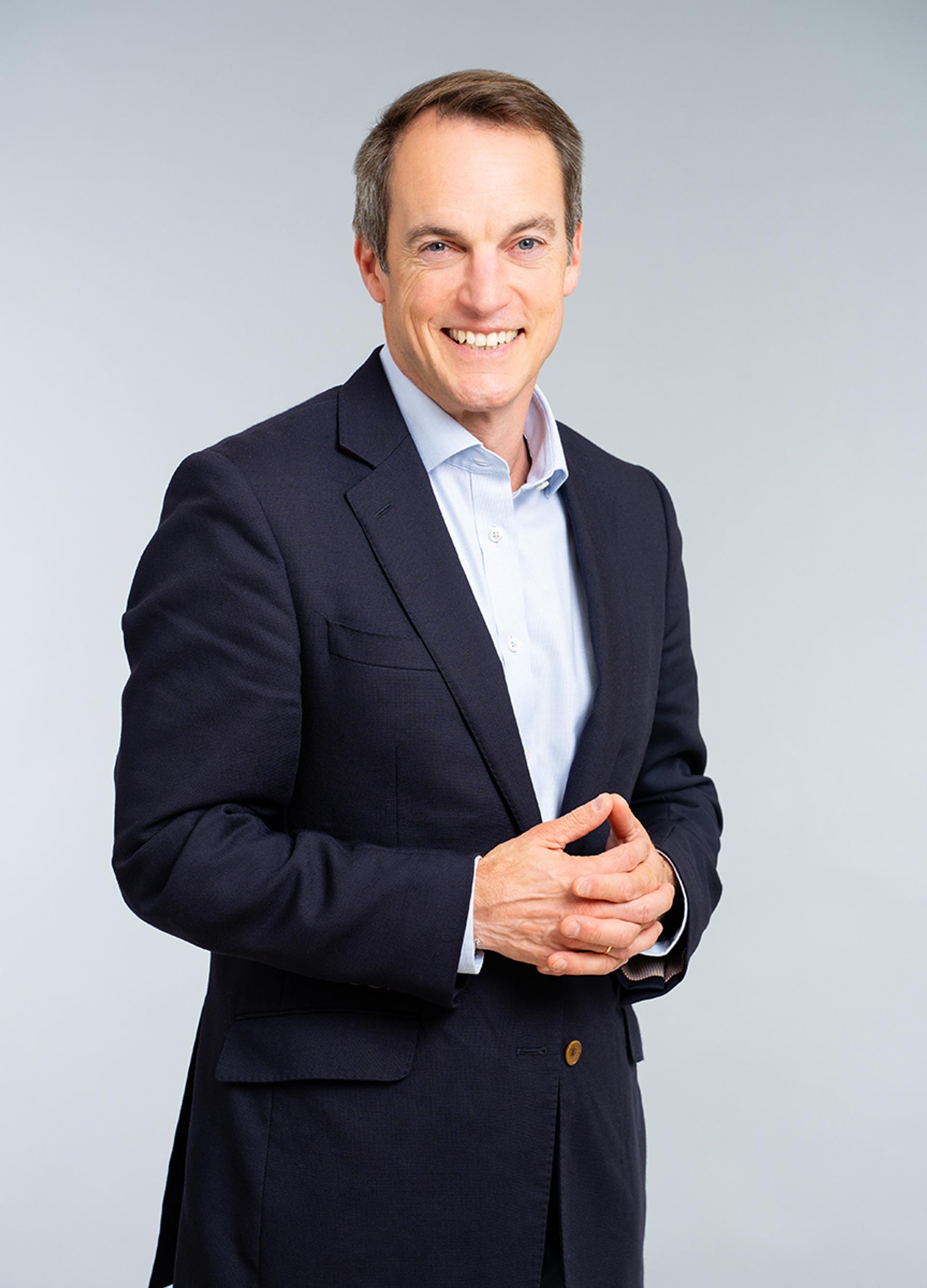 Portrait of a smiling man in a dark suit and light shirt standing against a plain grey background with hands clasped.