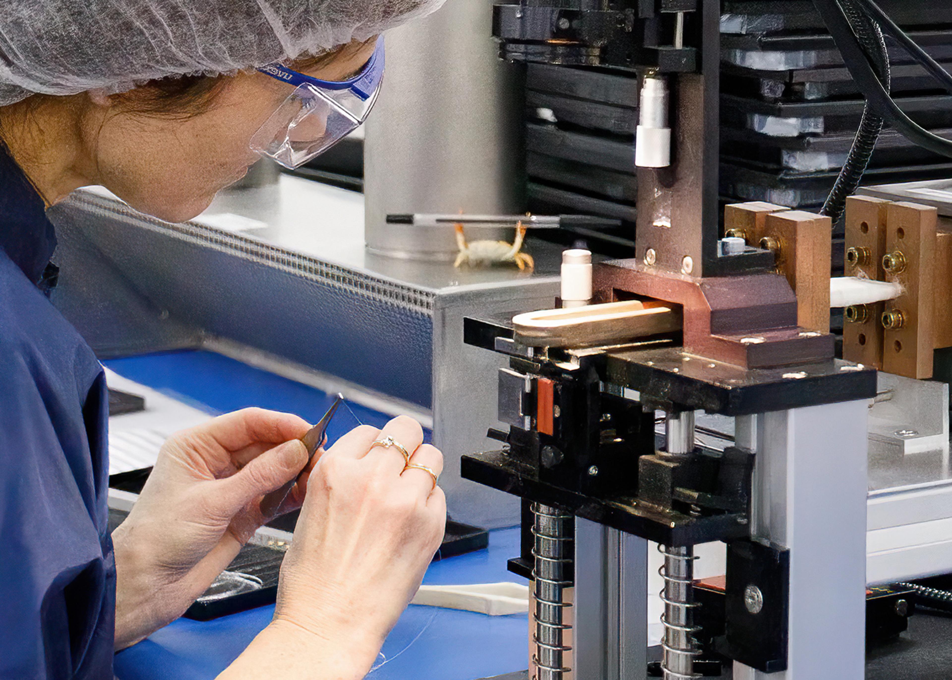 Technician wearing safety glasses and a hair cover precisely handling a fine fibre optic at a specialised assembly station, using precision tooling and equipment for fibre optic alignment and photonics component processing.