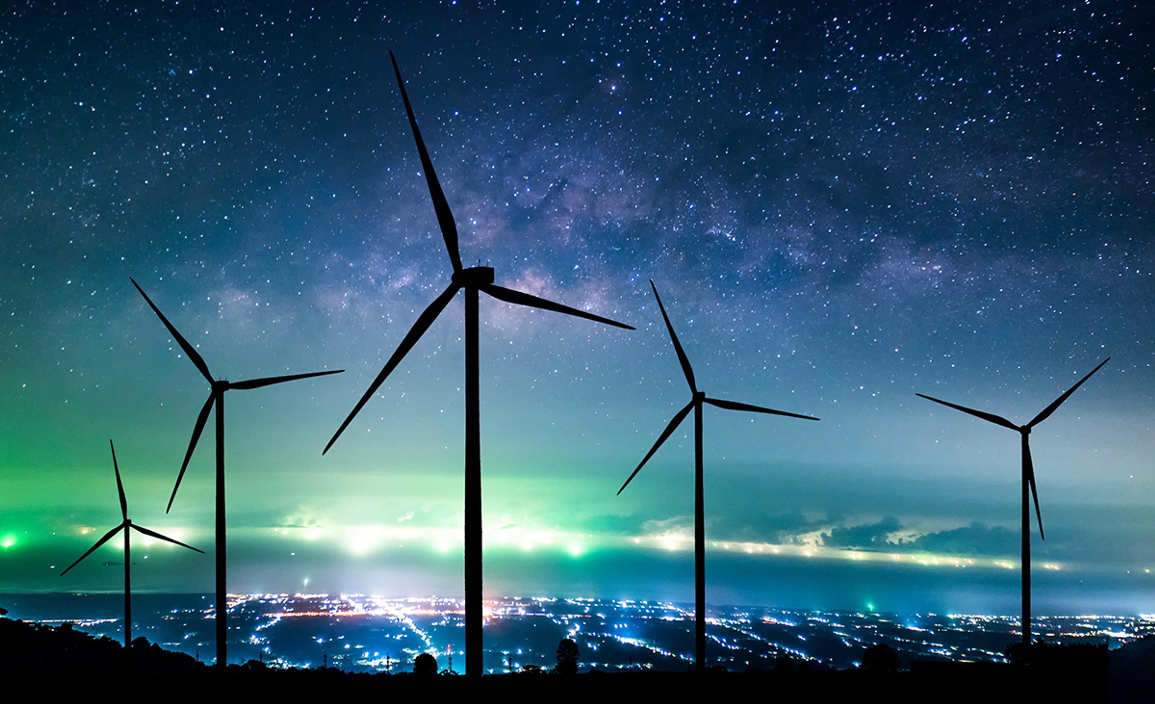 Silhouetted wind turbines at night overlooking a brightly lit cityscape, with the Milky Way visible across the sky.