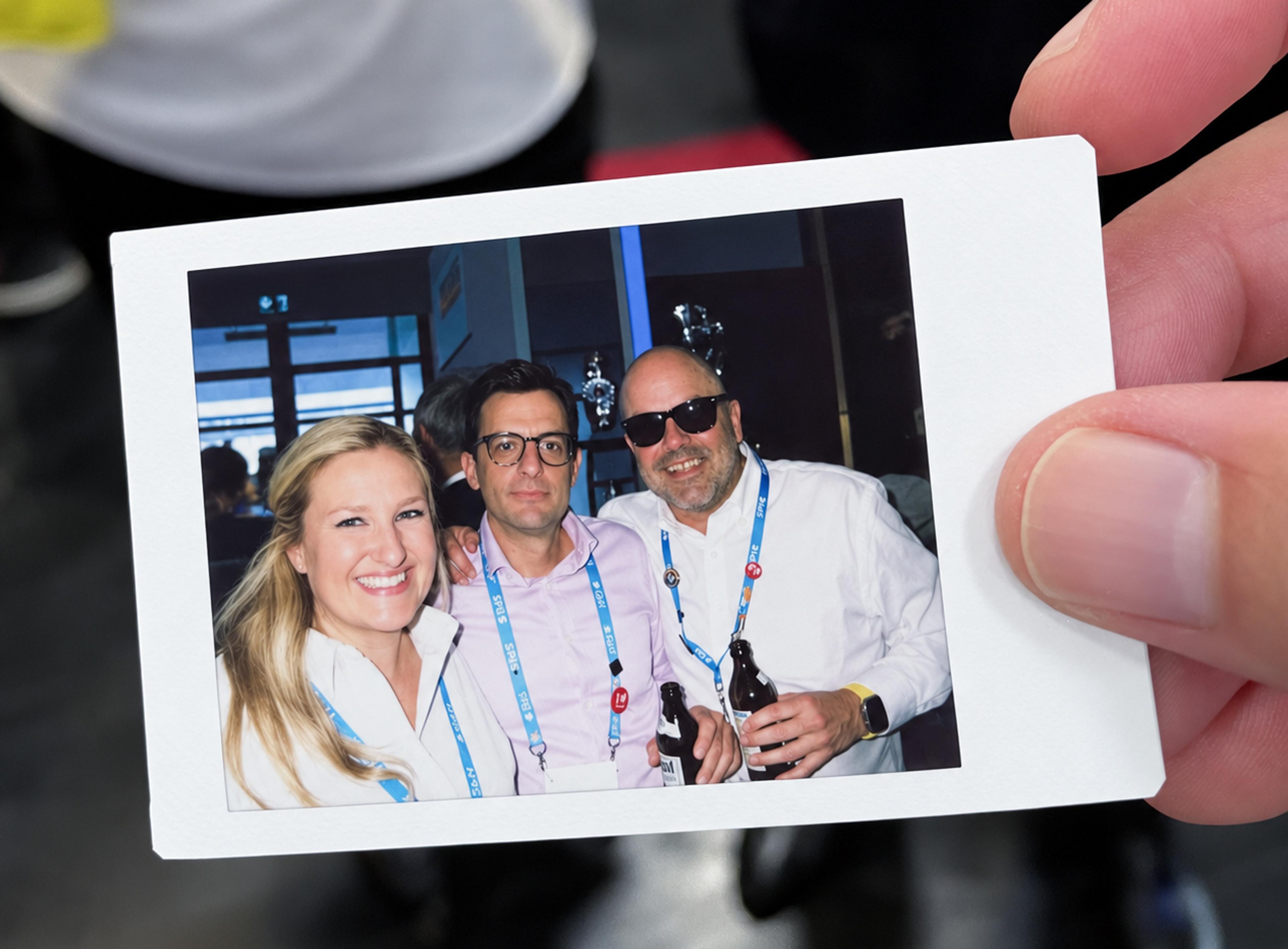 Hand holding a Polaroid photograph of three G&H team members posing together at an event, wearing G&H SPIE lanyards.